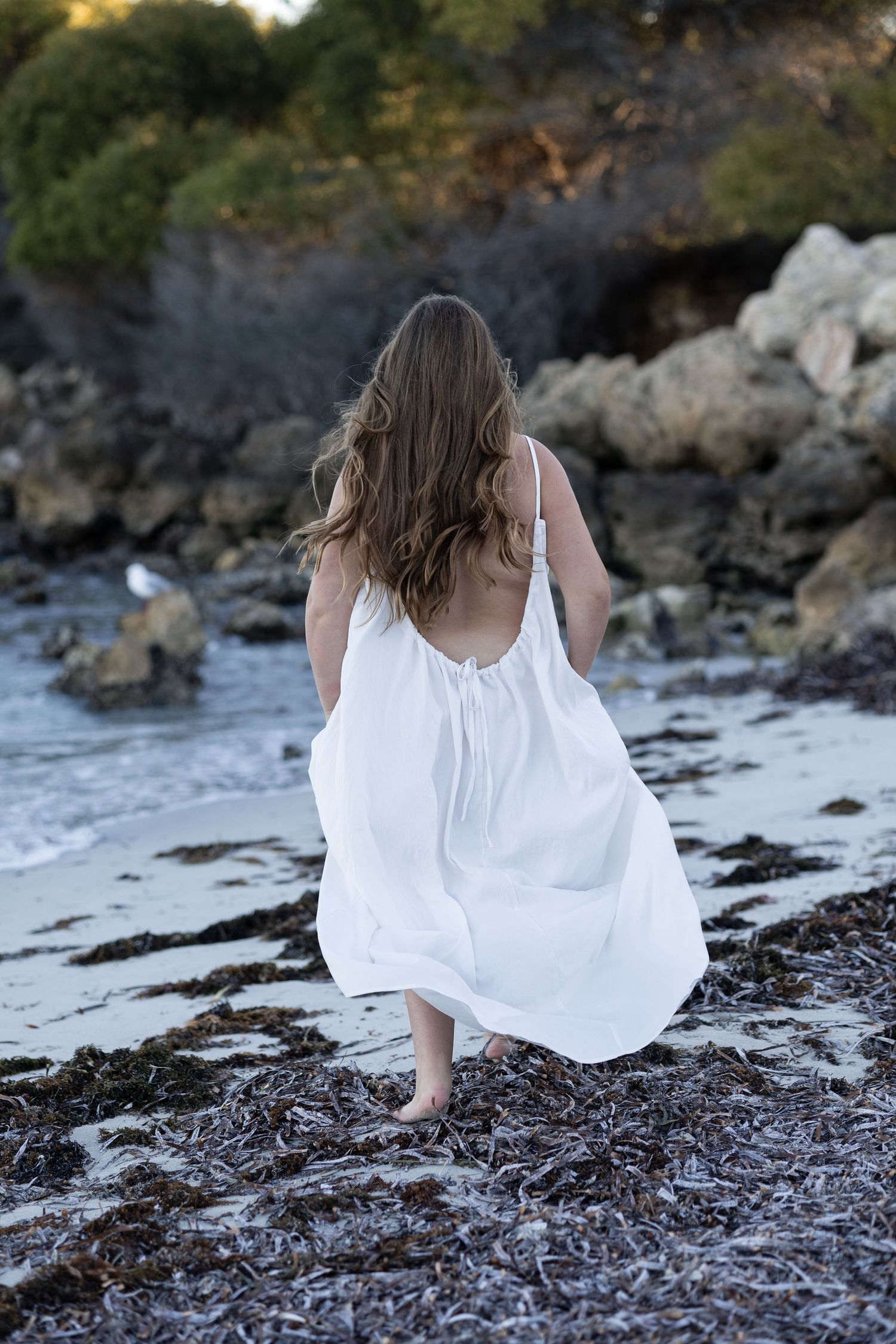 Woman in white sundress walking barefoot on rocky beach with flowing hair and ocean waves.