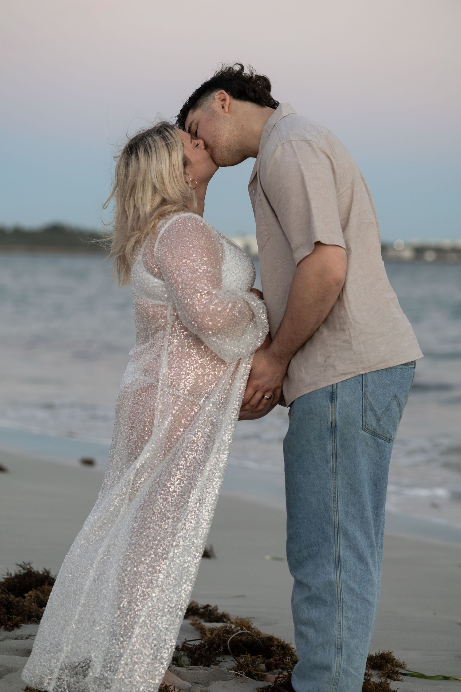 Couple shares a kiss on the beach, woman in sparkly white maternity gown, man in beige shirt and jeans.