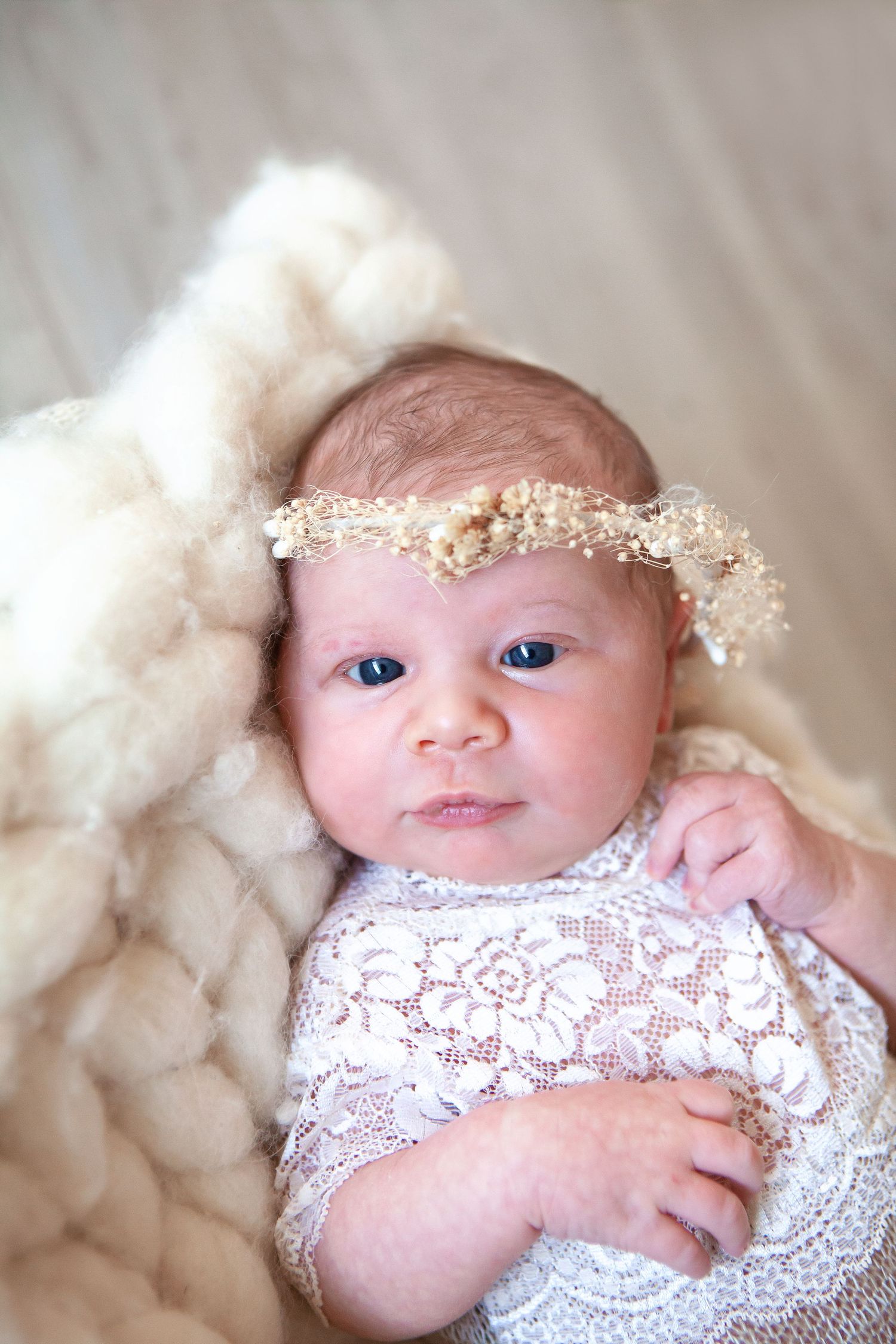 Alert newborn baby in white lace outfit wearing a gold floral crown on a wool blanket.