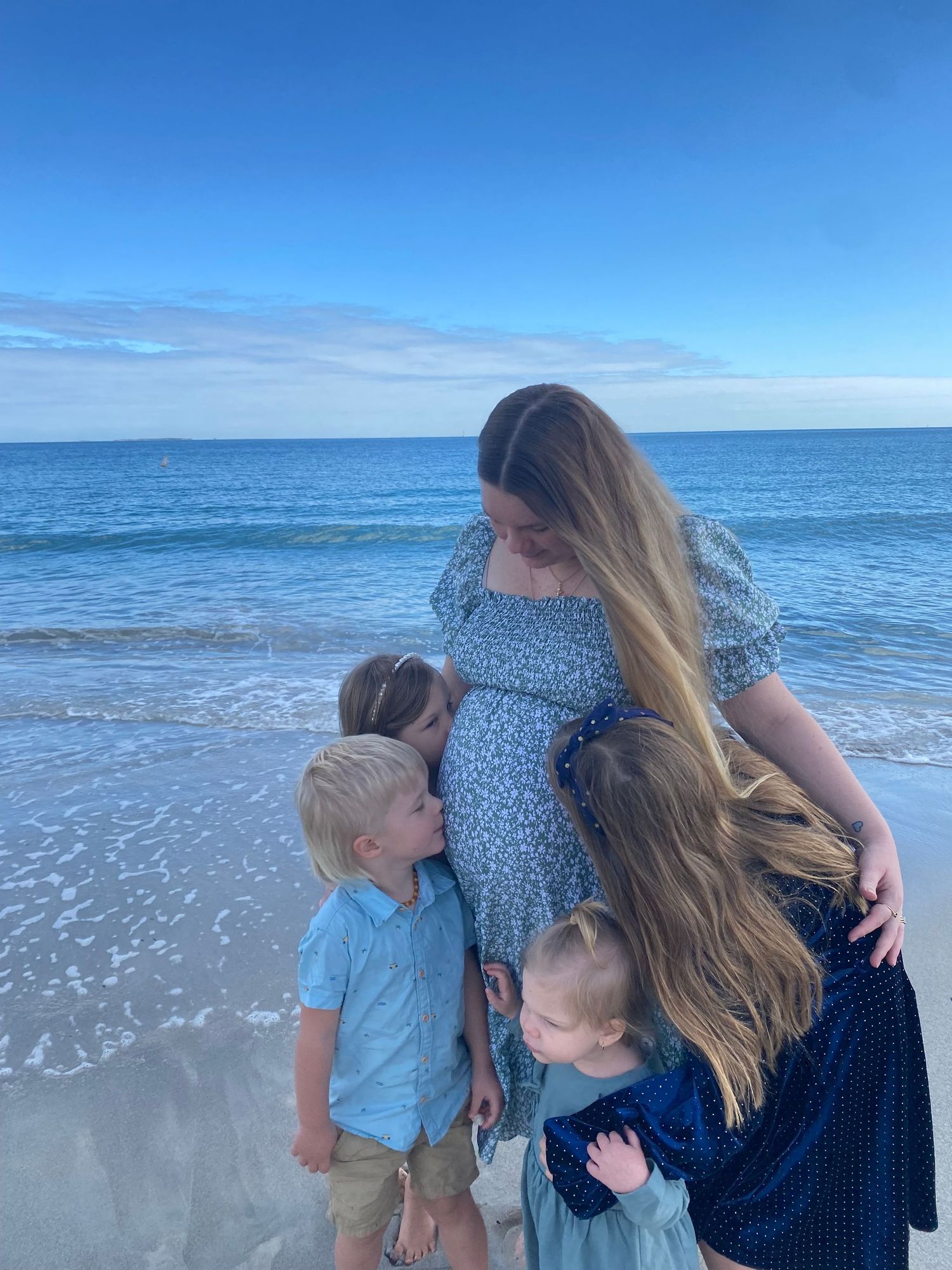 Pregnant woman in floral dress surrounded by three young children on sandy beach with blue ocean.