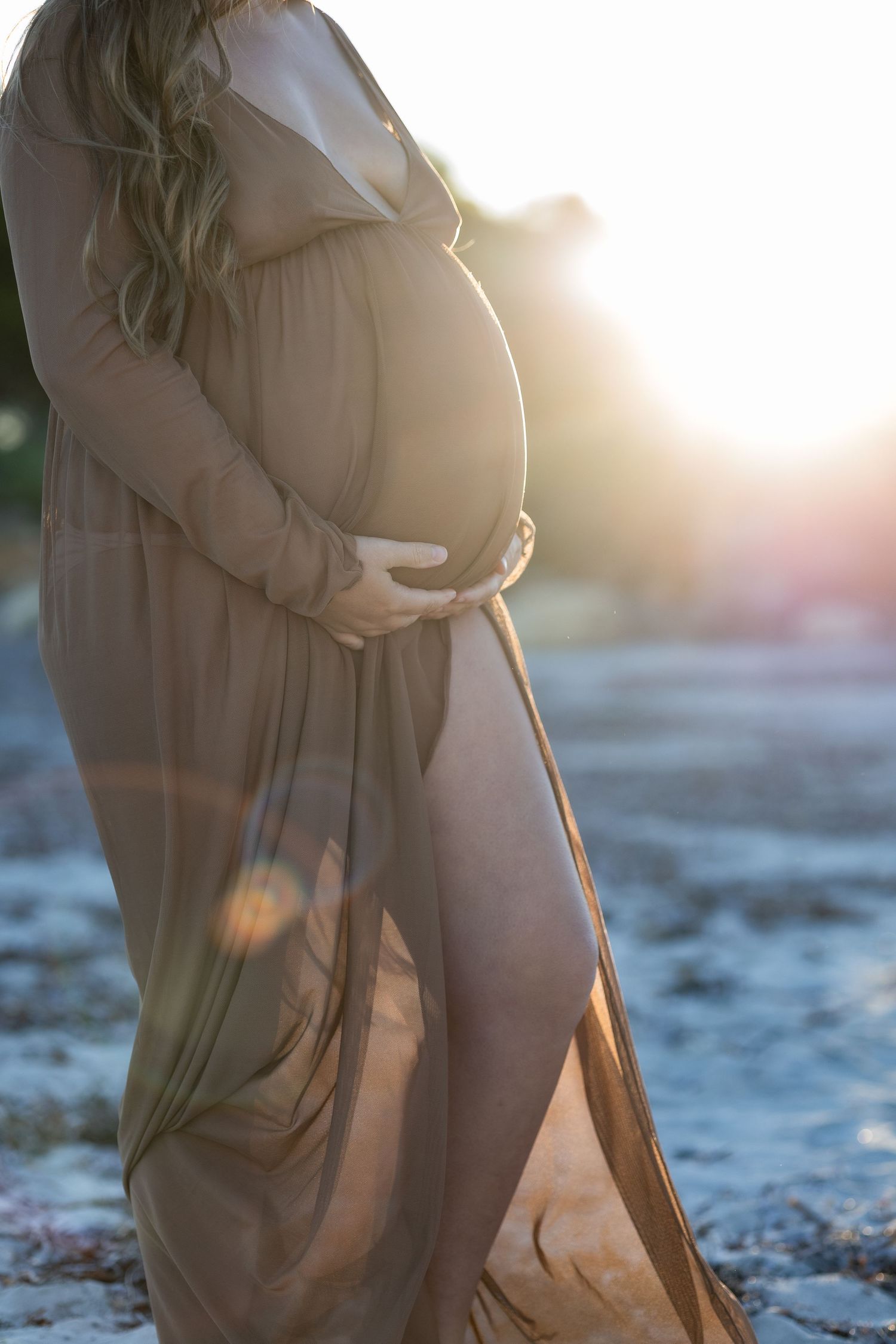 Pregnant woman in sheer olive maxi dress cradling baby bump outdoors in golden sunset light.