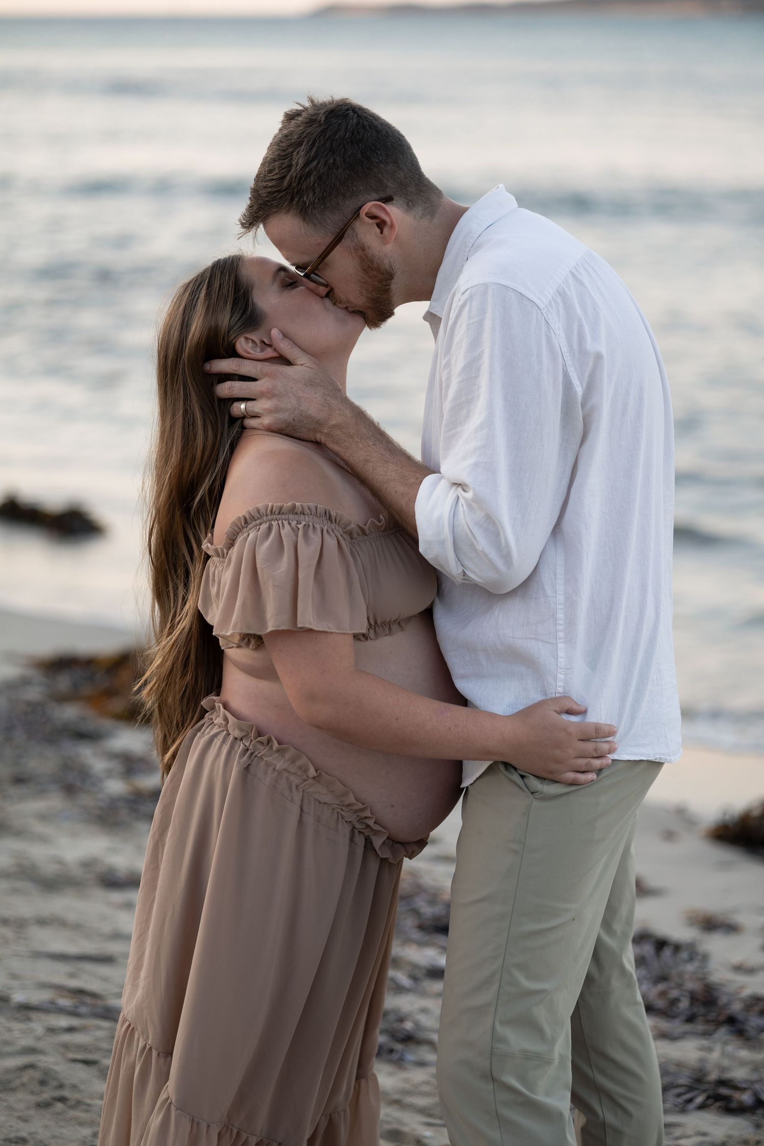 Pregnant couple sharing a romantic kiss on a beach at sunset, woman in tan off-shoulder dress.