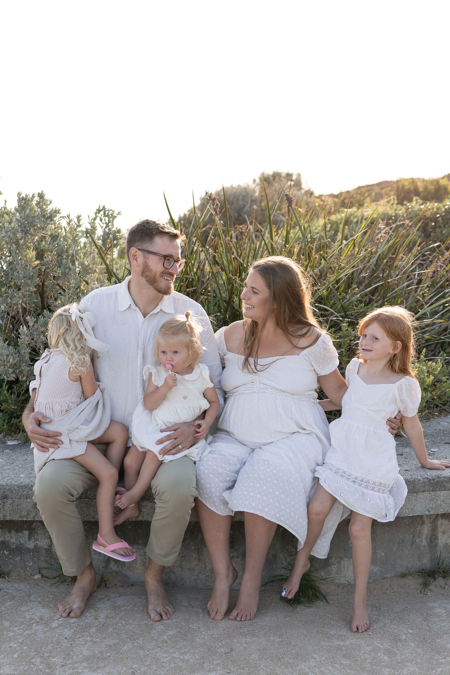 A happy family of five sits on a stone bench outdoors, with three young girls and a pregnant mother in white dress.  Cape Peron Perth