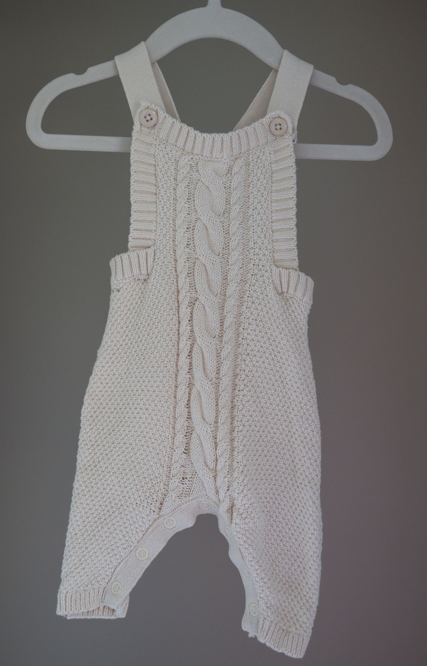 White cable-knit baby romper with button straps displayed on a white hanger against a gray background. Taken by Golden Hour Perth