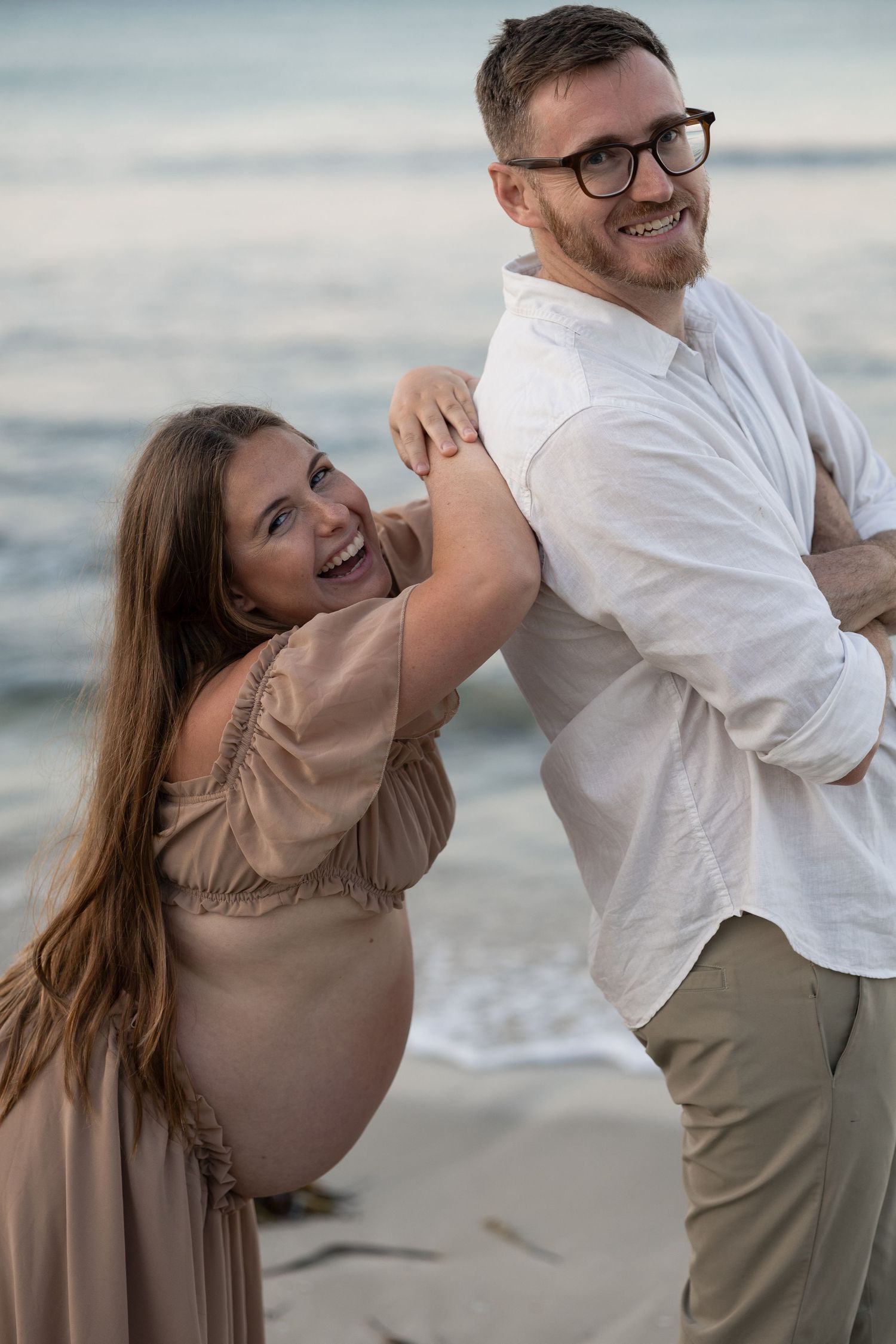 Smiling pregnant couple posing playfully on a sandy beach at sunset, woman in beige dress hugging man in white shirt.