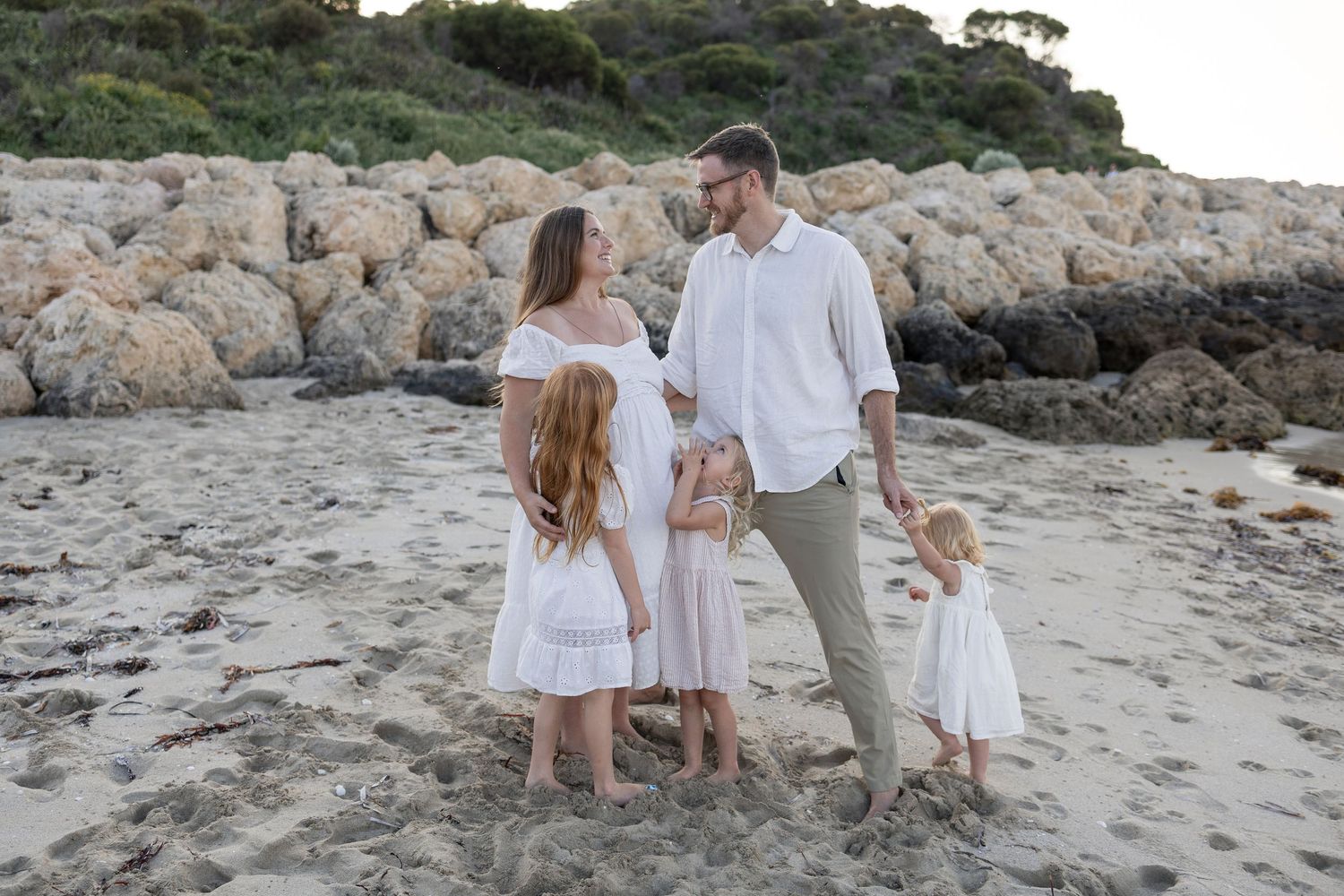 Family of four in white outfits posing on a sandy beach with rocky coastline backdrop at golden hour. Cape Peron Perth