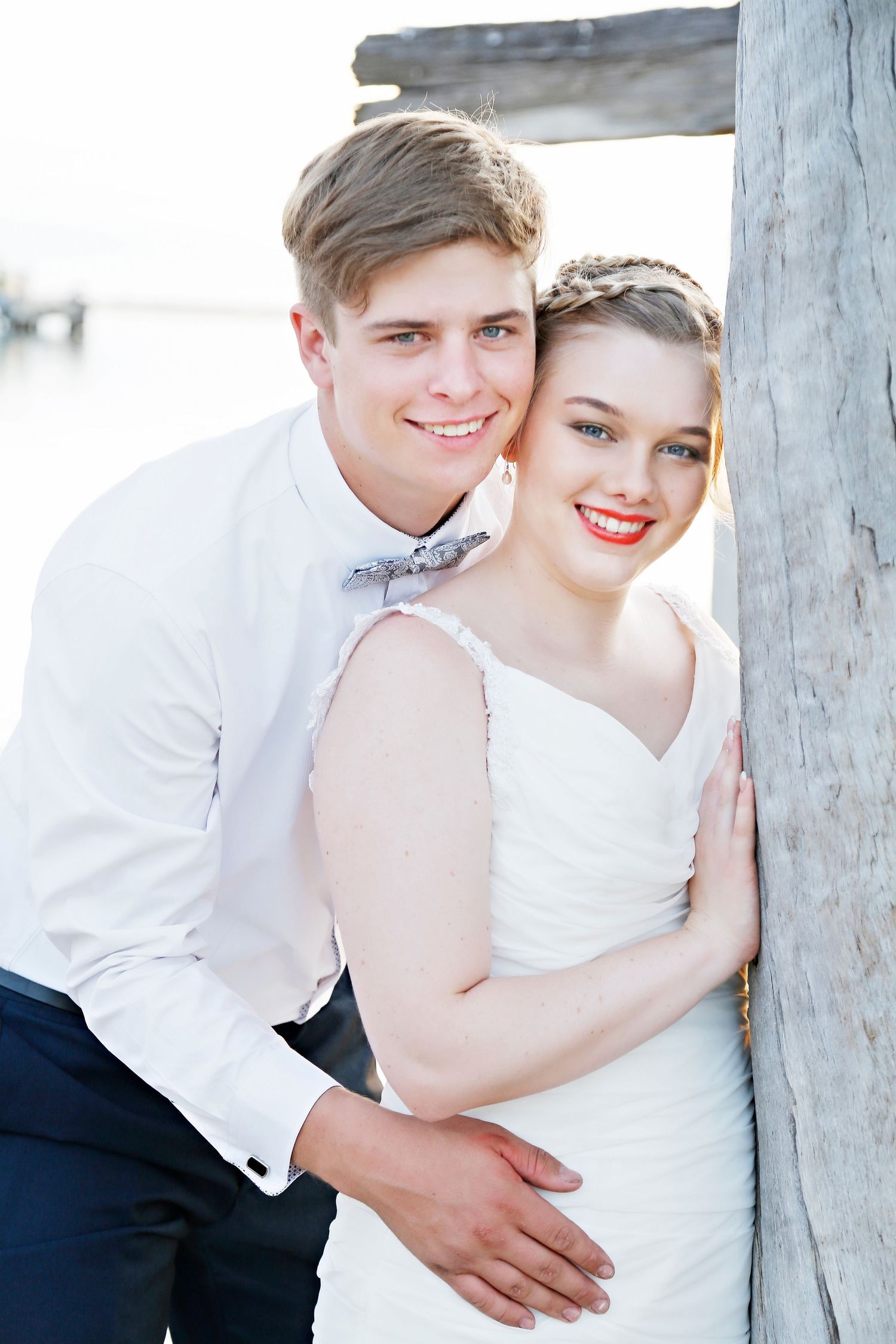 Happy newlywed couple posing near wooden pier by water, groom in white shirt and bow tie with bride in white dress.