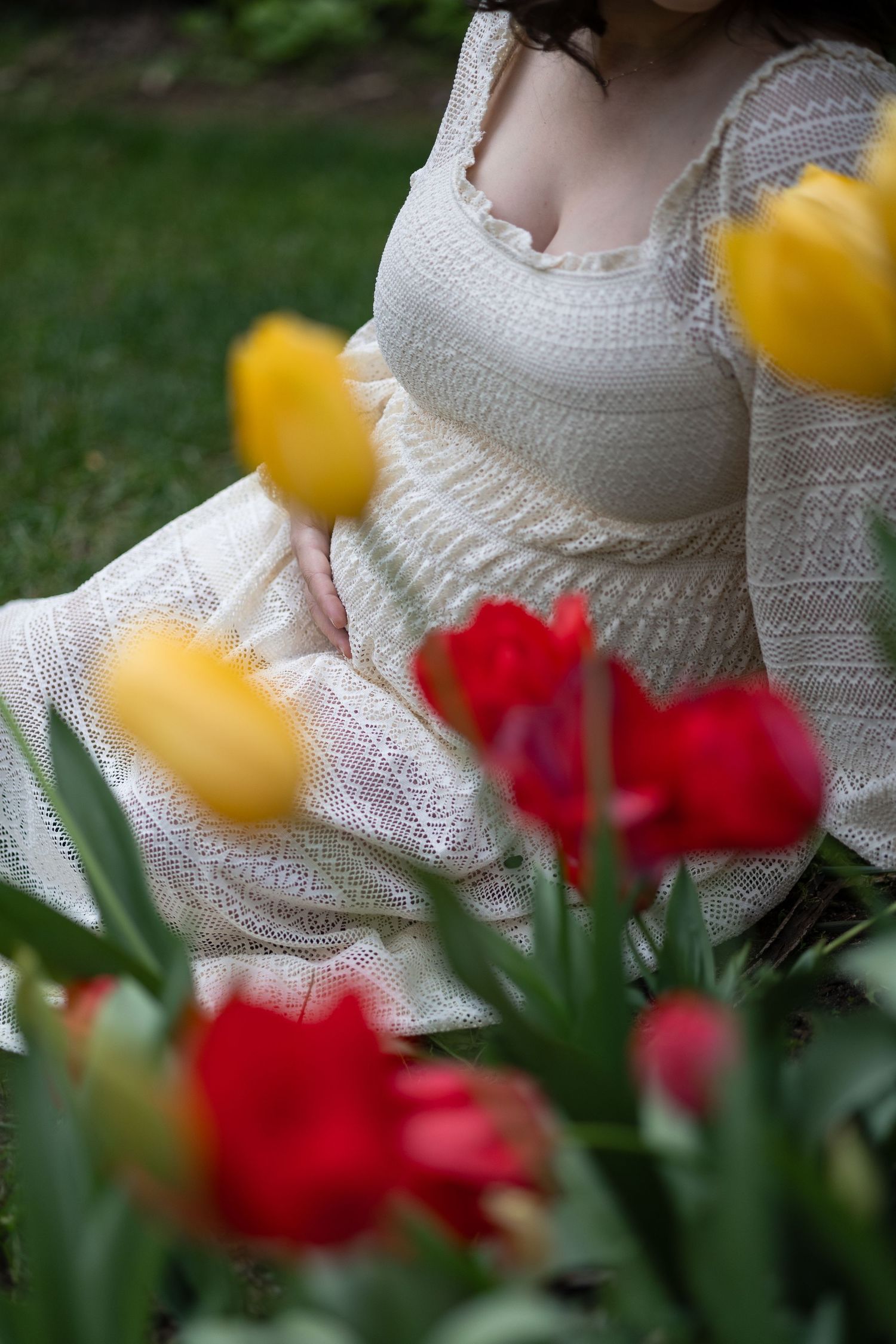 Pregnant woman in white lace dress sitting among red and yellow tulips in a garden.