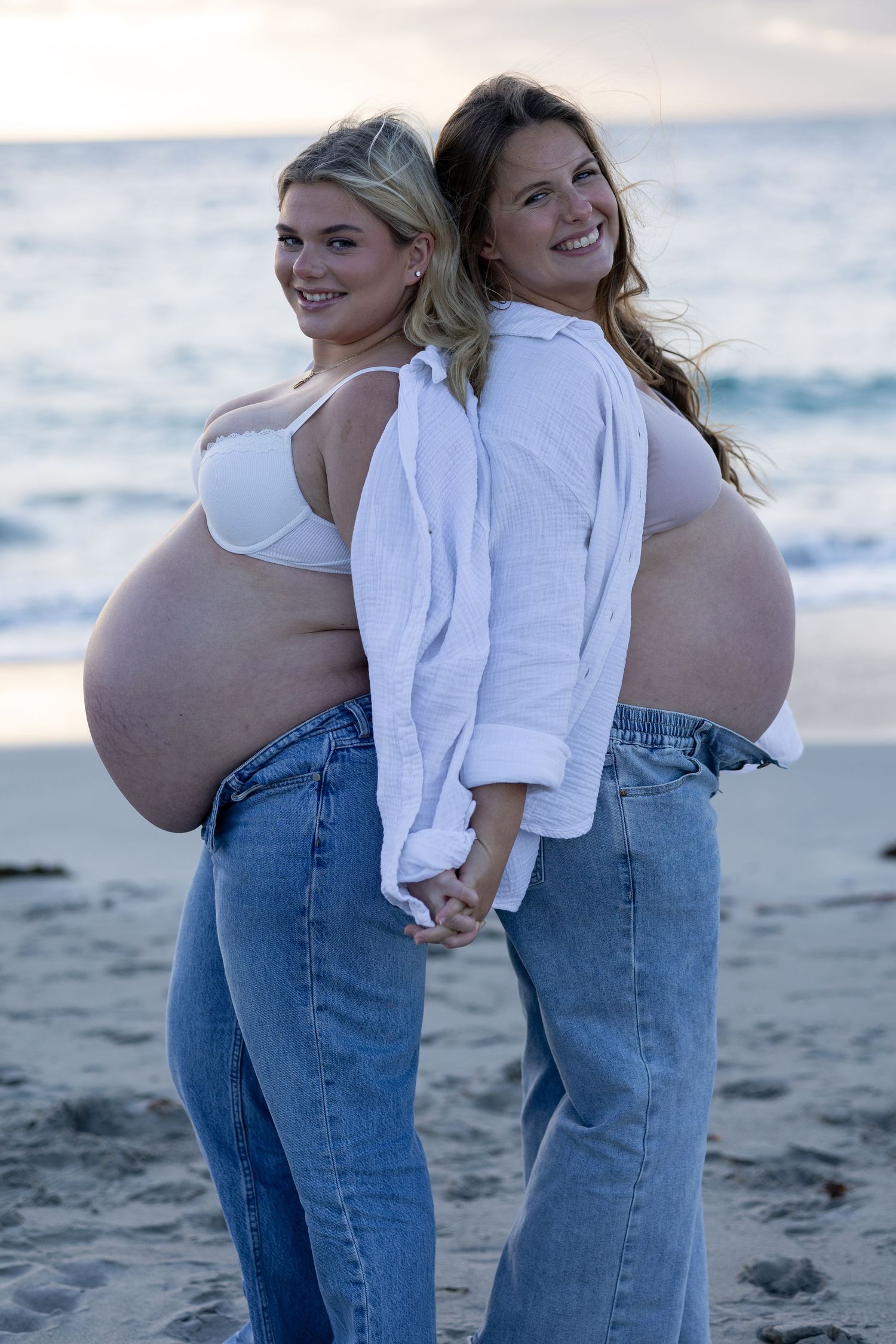 Two pregnant women in jeans pose back-to-back on a beach at sunset, smiling and holding hands.