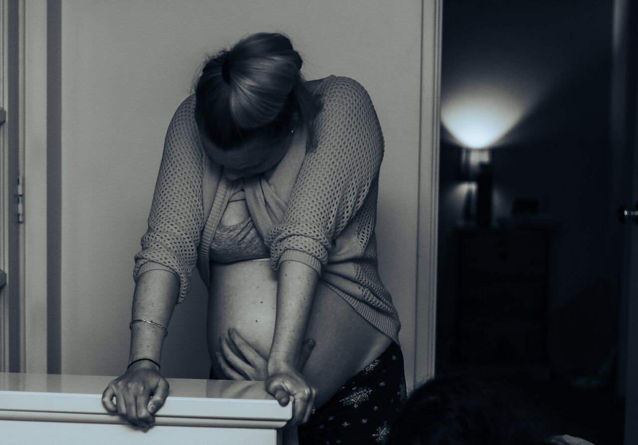 Black and white photo of a distressed woman hunched over a windowsill in a dimly lit room.
