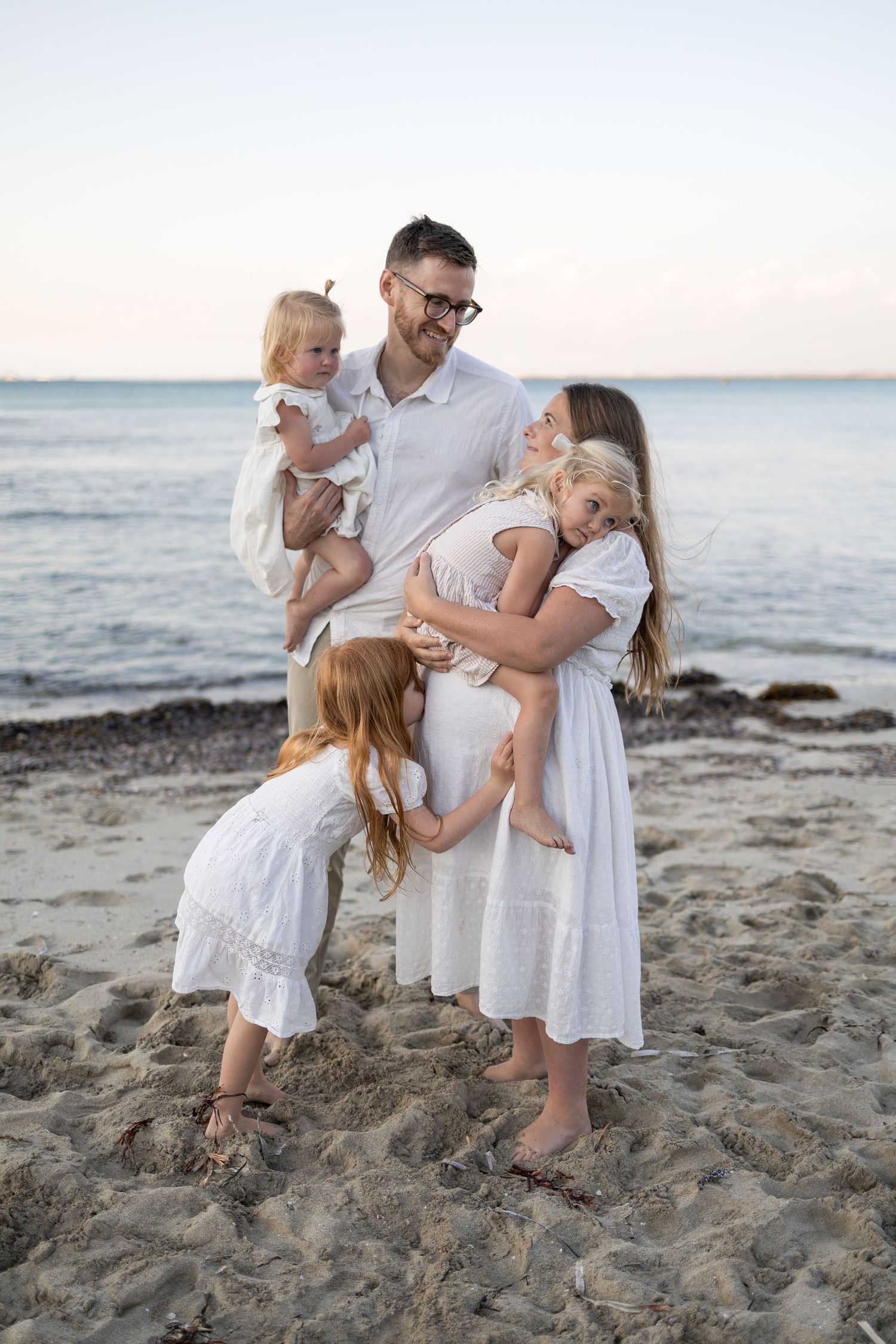 Family of five in white outfits posing on sandy beach at sunset, father holding toddler while children embrace pregnant mother.