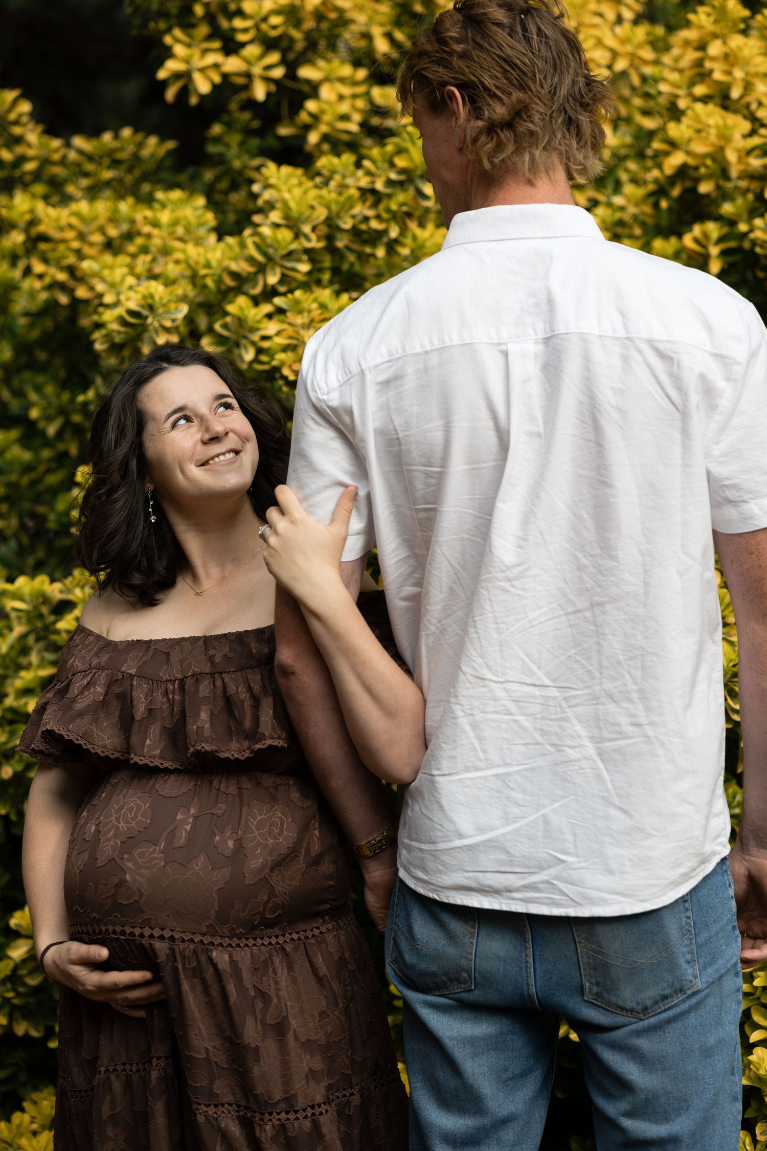 Pregnant woman in brown dress smiles at partner in white shirt outdoors near yellow-green foliage.  Araluen Perth