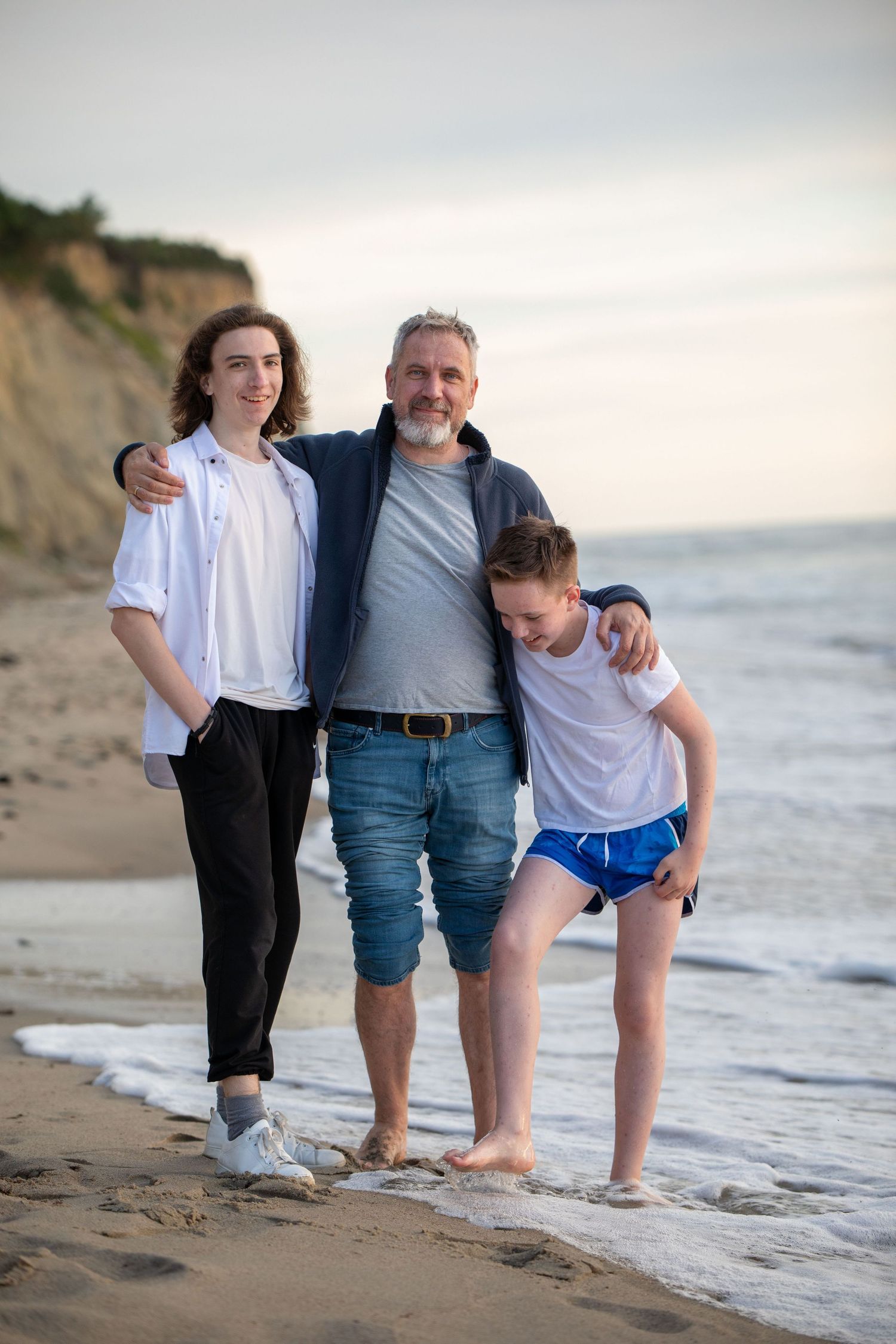 Father with two teenage children embracing on a sandy beach at sunset.