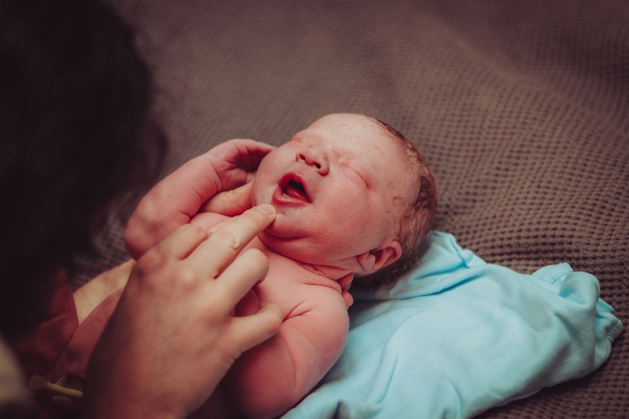 Newborn baby crying while being held by mother's hands on a soft gray blanket.