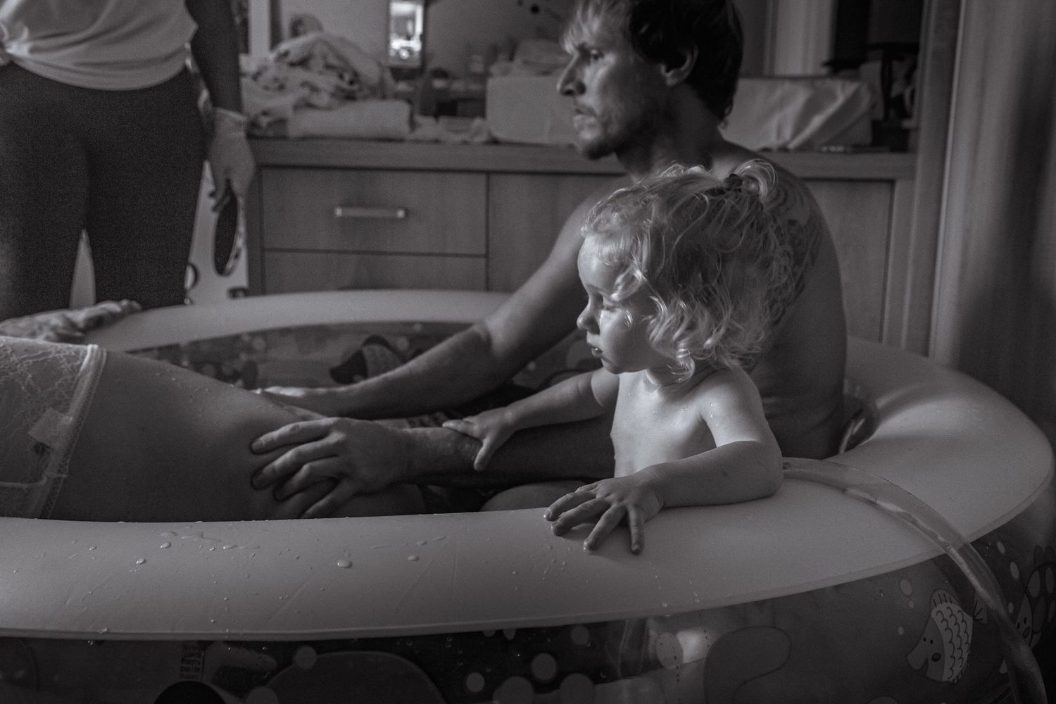 Black and white photo of pregnant woman in birth pool with toddler during home water birth.