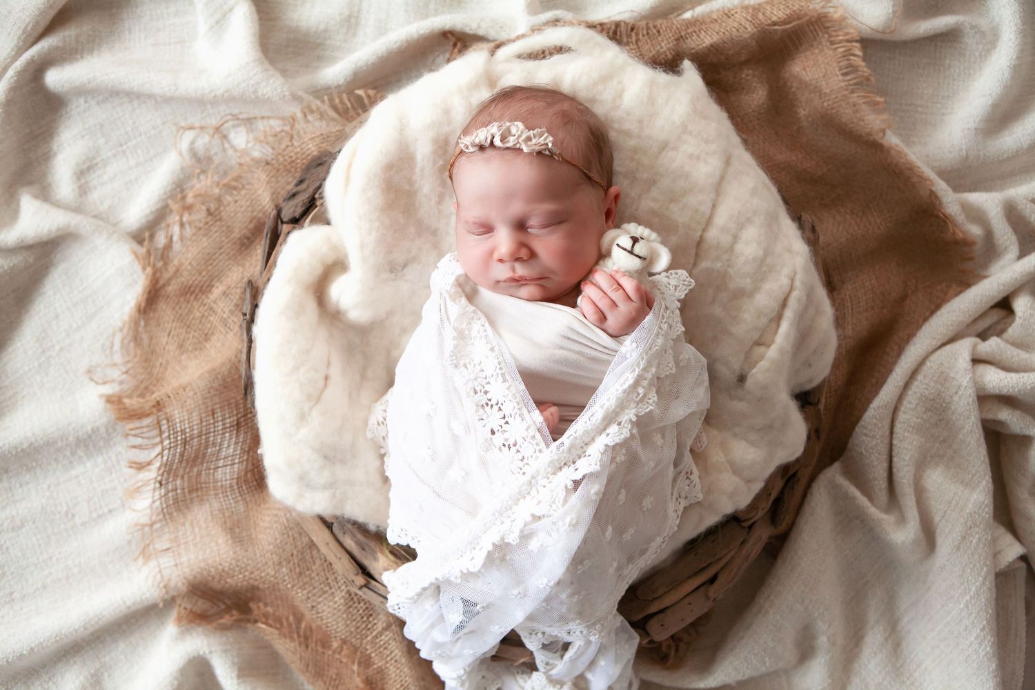 Newborn baby swaddled in white lace wrap with floral headband lying in a cozy basket.