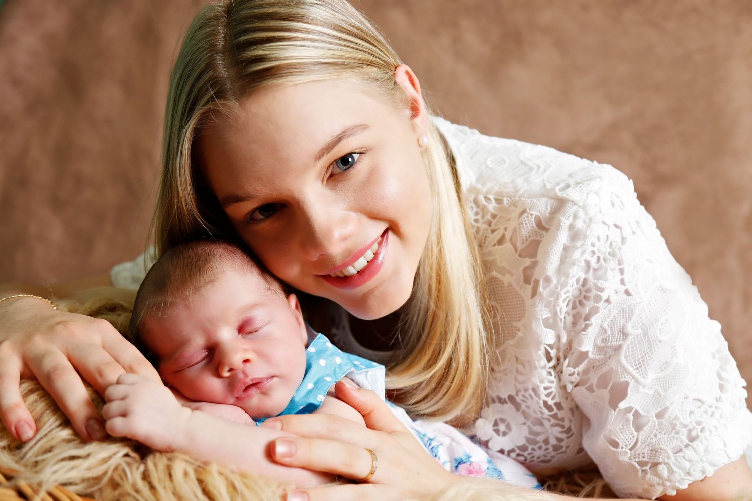 Smiling blonde mother in white lace dress posing with sleeping newborn baby dressed in blue onesie.