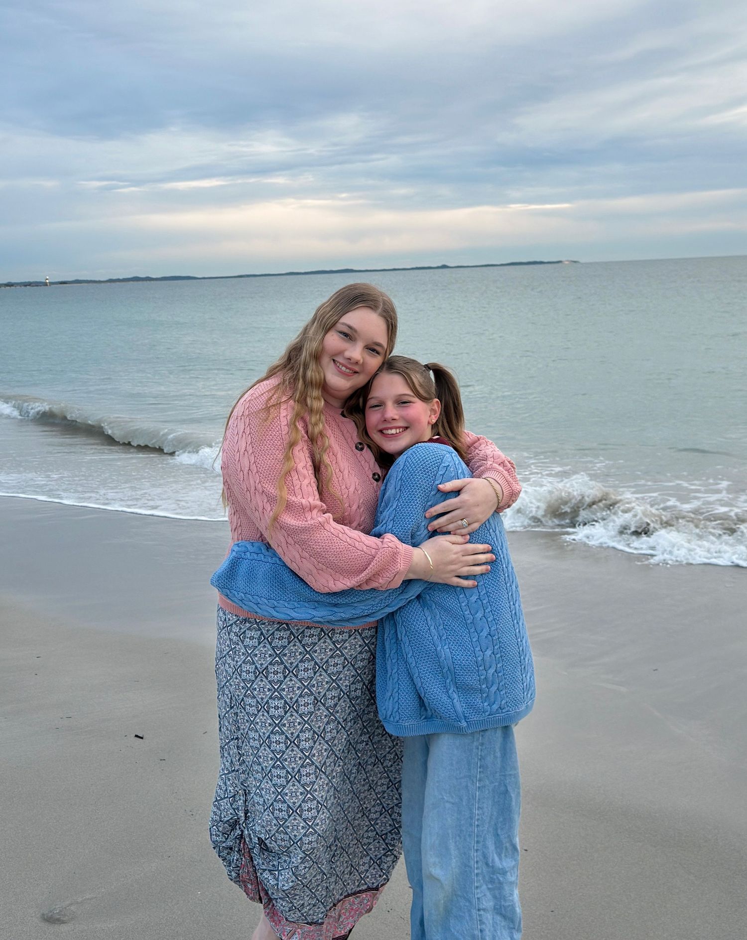 Two girls hugging and smiling on a beach, one in pink sweater and one in blue, with ocean waves behind them.