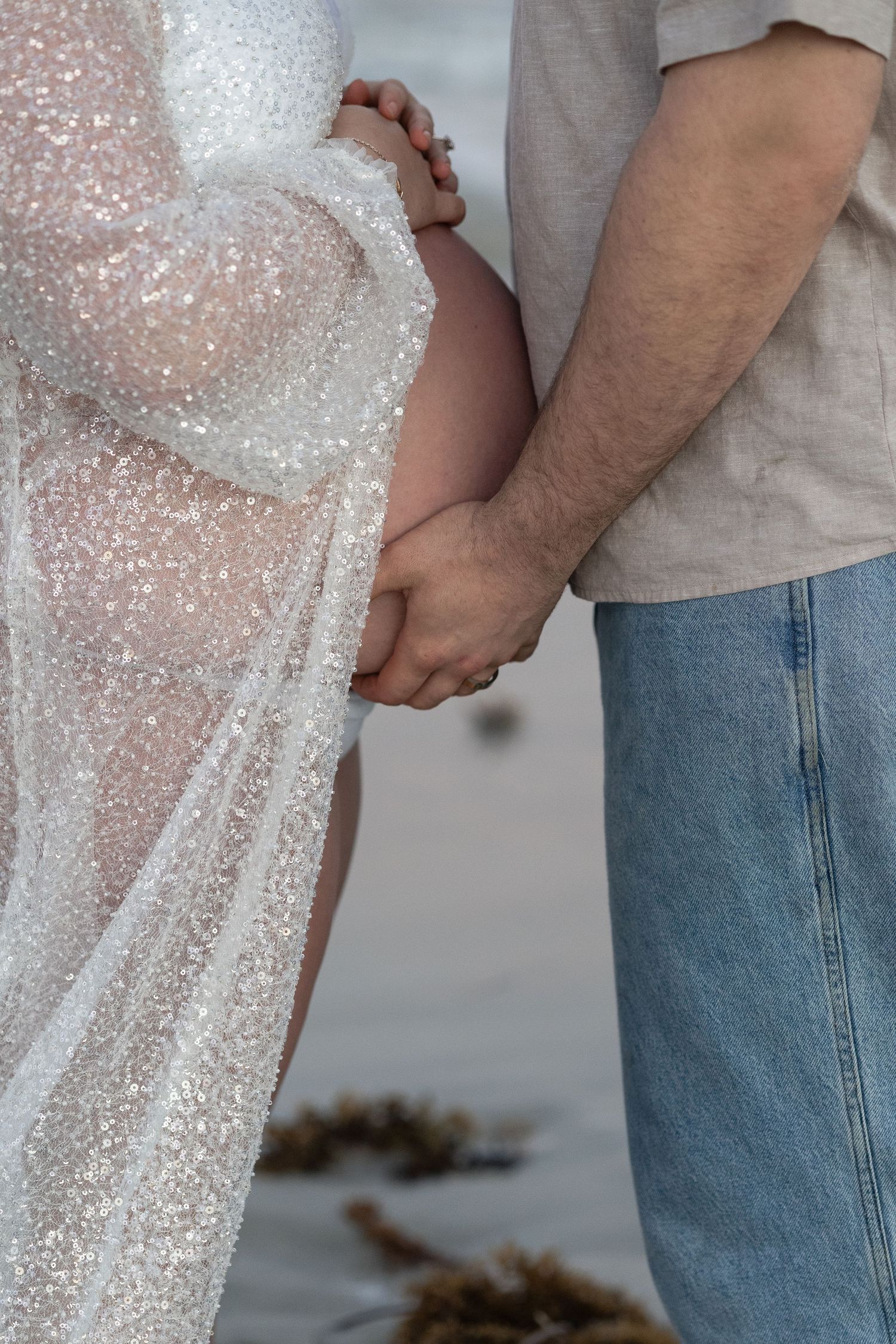 Pregnant woman in sparkly white gown holds hands with partner in jeans at beach maternity photoshoot.