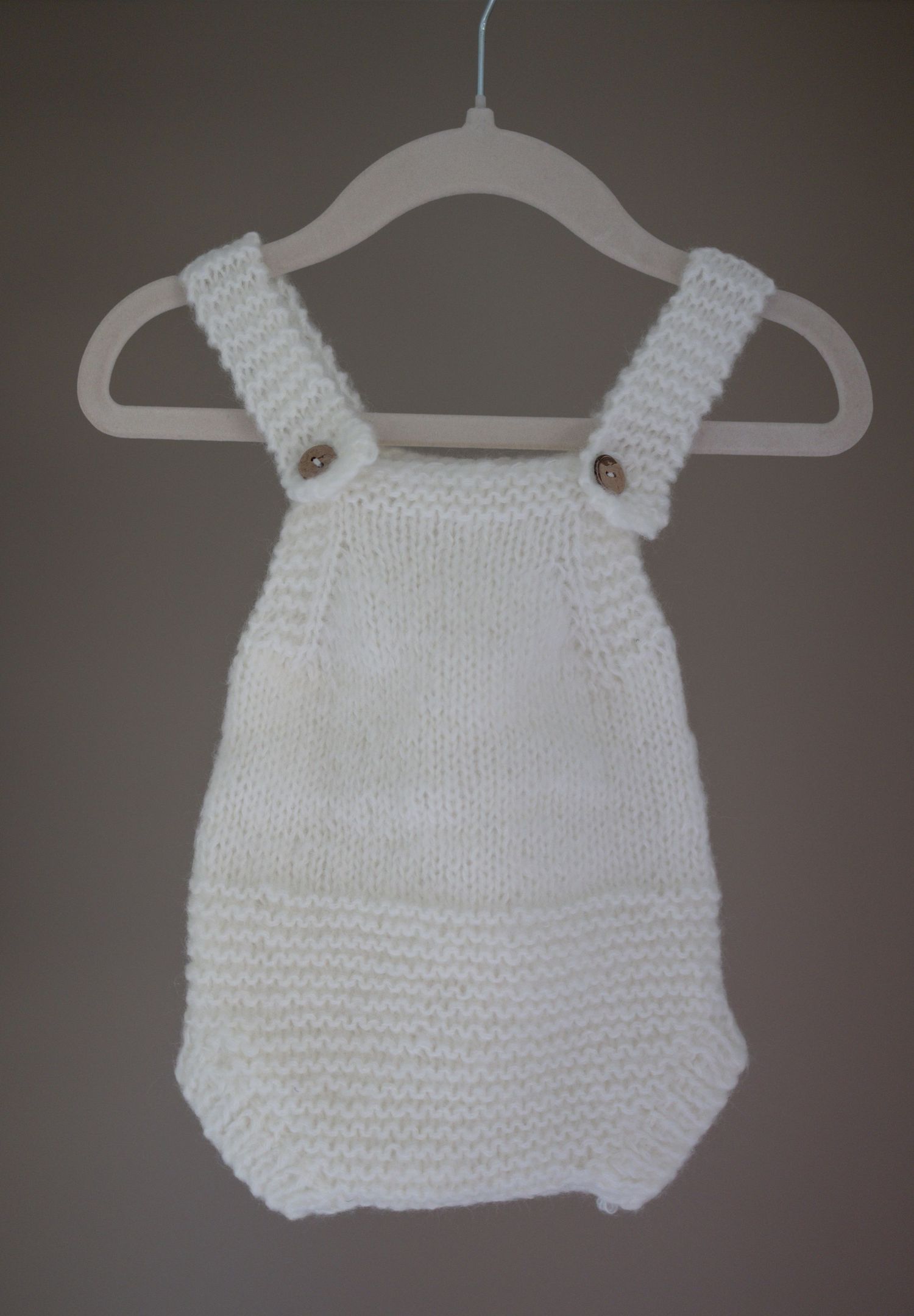 White hand-knitted baby romper with shoulder straps and buttons, displayed on a small white hanger. Taken by Golden Hour Perth