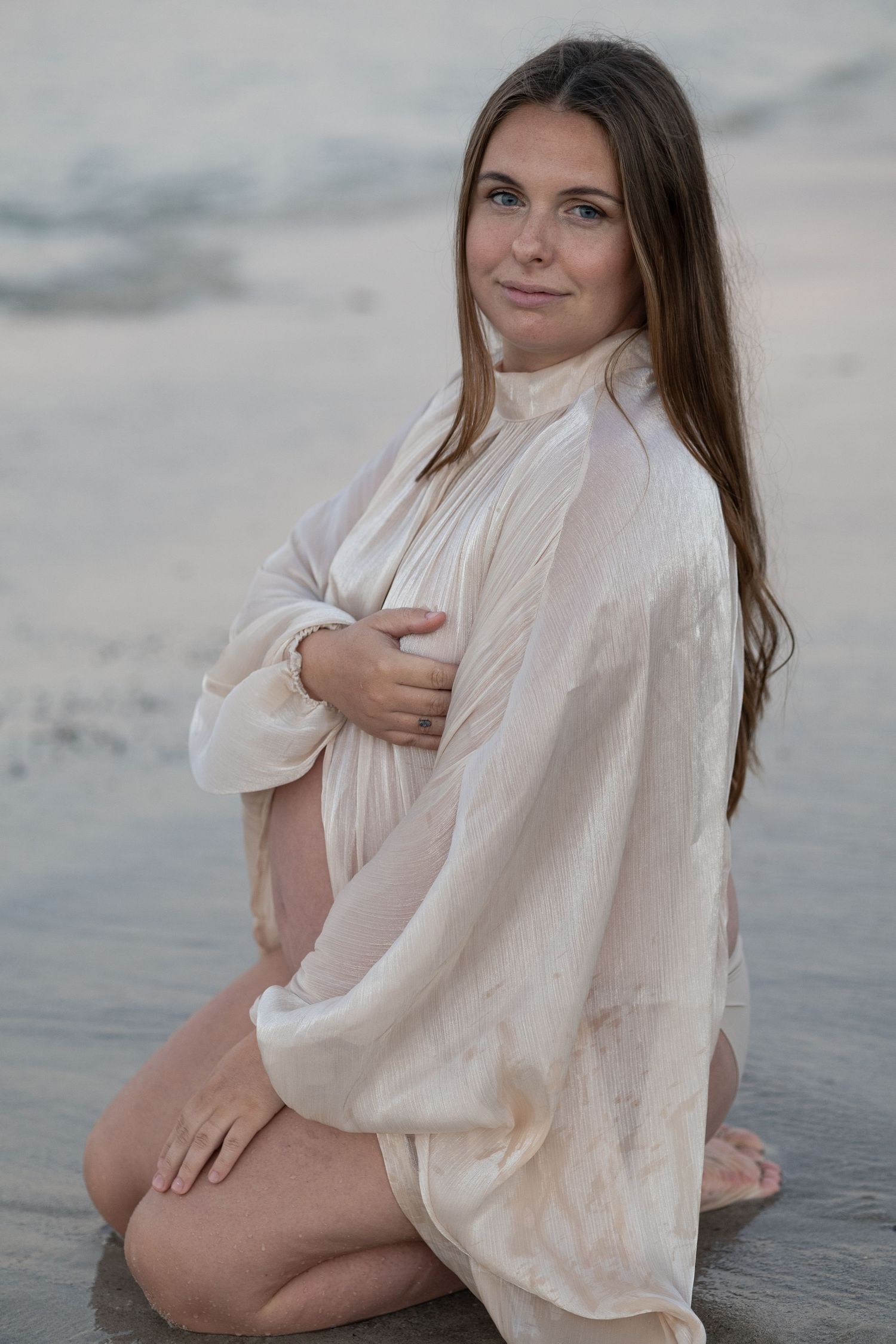 Pregnant woman in white sheer robe sitting on beach shore, cradling baby bump, with ocean waves in background. Taken by Golden Hour Perth