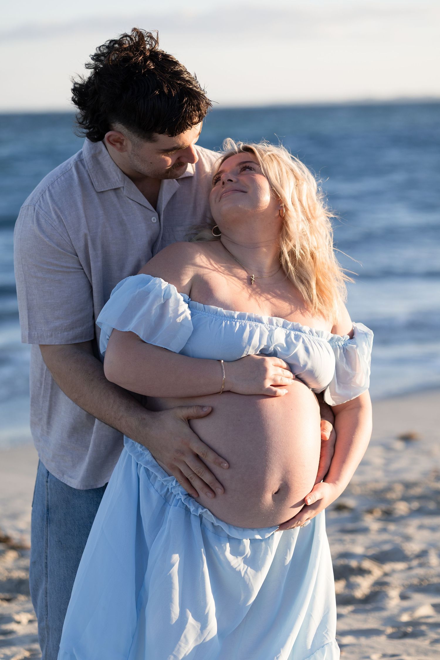 Couple on beach, man embracing pregnant woman in blue dress, sharing tender moment by the ocean at sunset. Woodman Point Perth