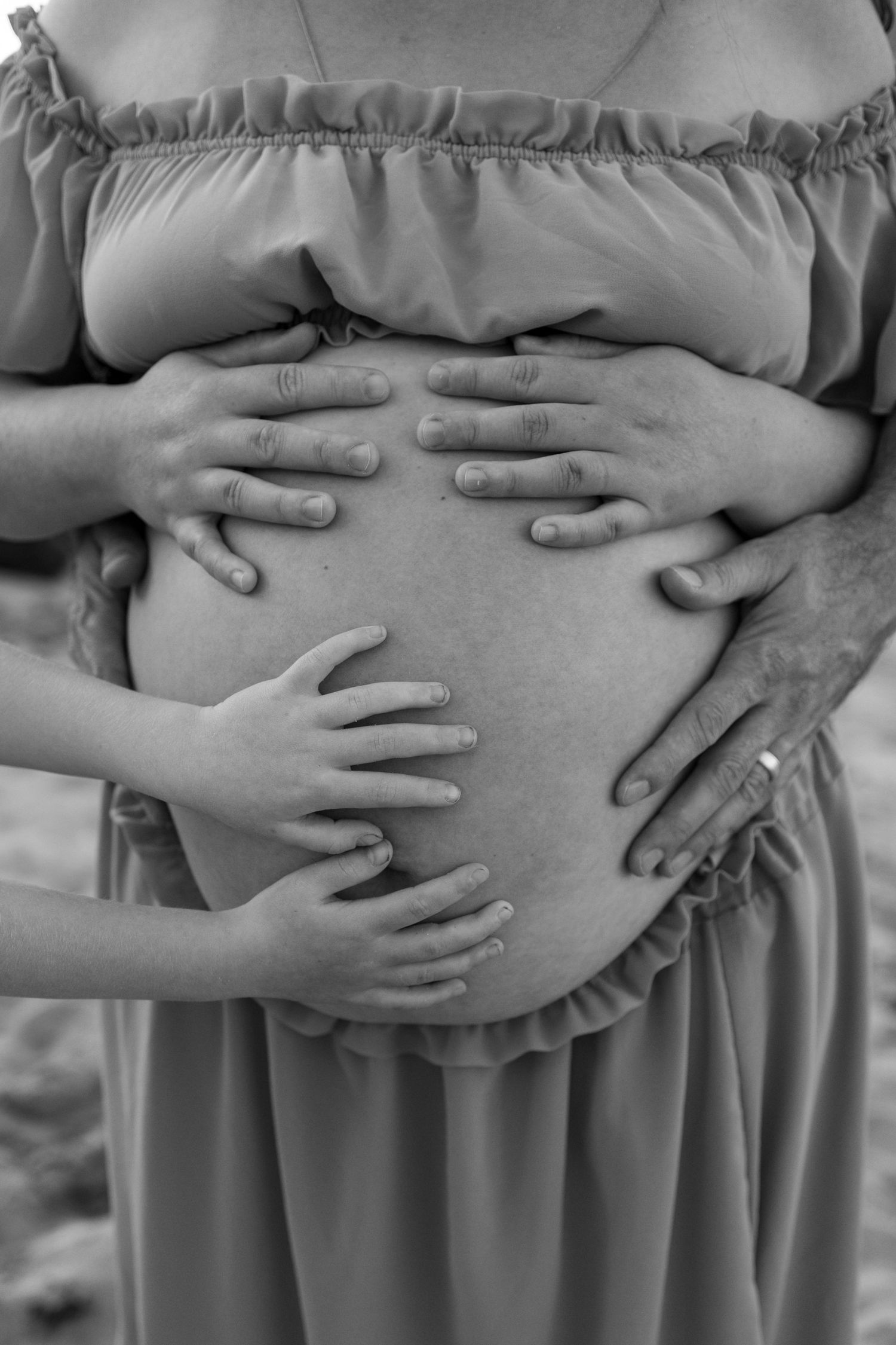 Black and white photo of pregnant woman's belly being cradled by multiple hands, including a child's.