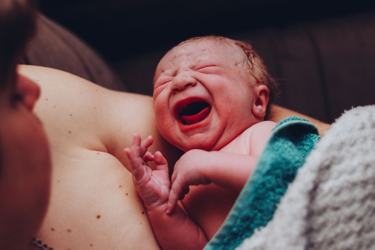 Newborn baby crying moments after birth, held against mother's chest, wrapped in teal towel.