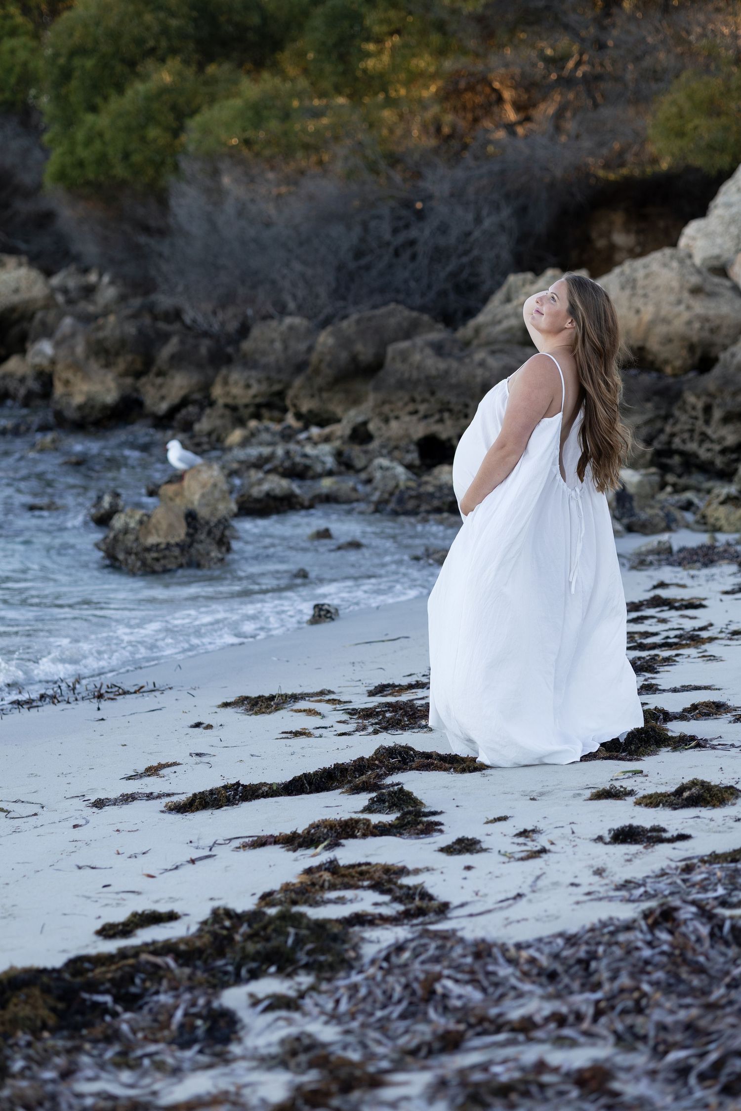 Pregnant woman in flowing white dress stands on seaweed-covered beach gazing upward near rocky cliffs.