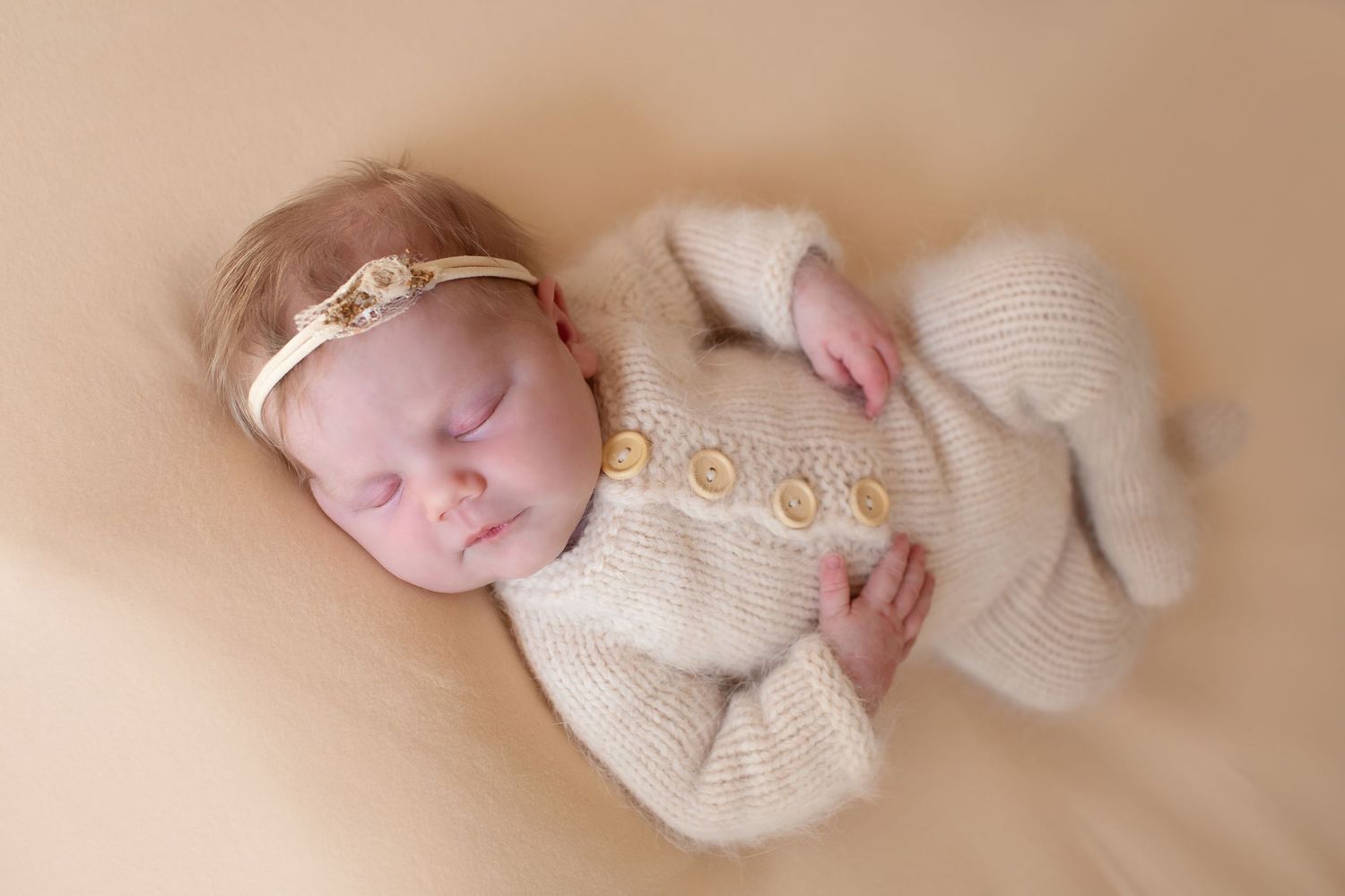 Sleeping newborn baby wearing a cream knit romper and gold headband on beige backdrop.
