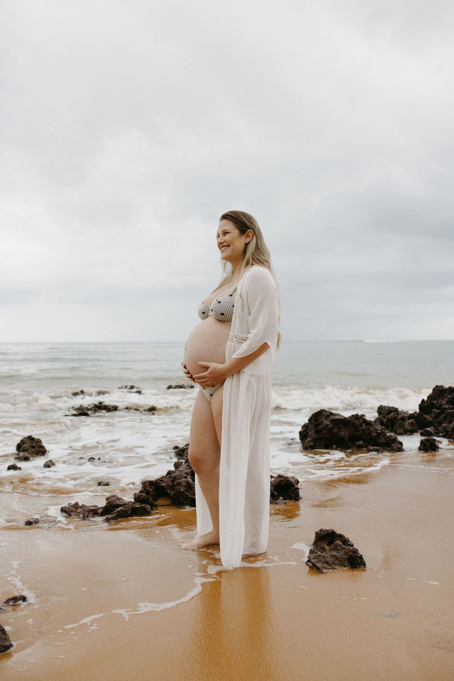 Smiling pregnant woman in bikini and sheer white cover-up cradling her bump on a rocky sandy beach.