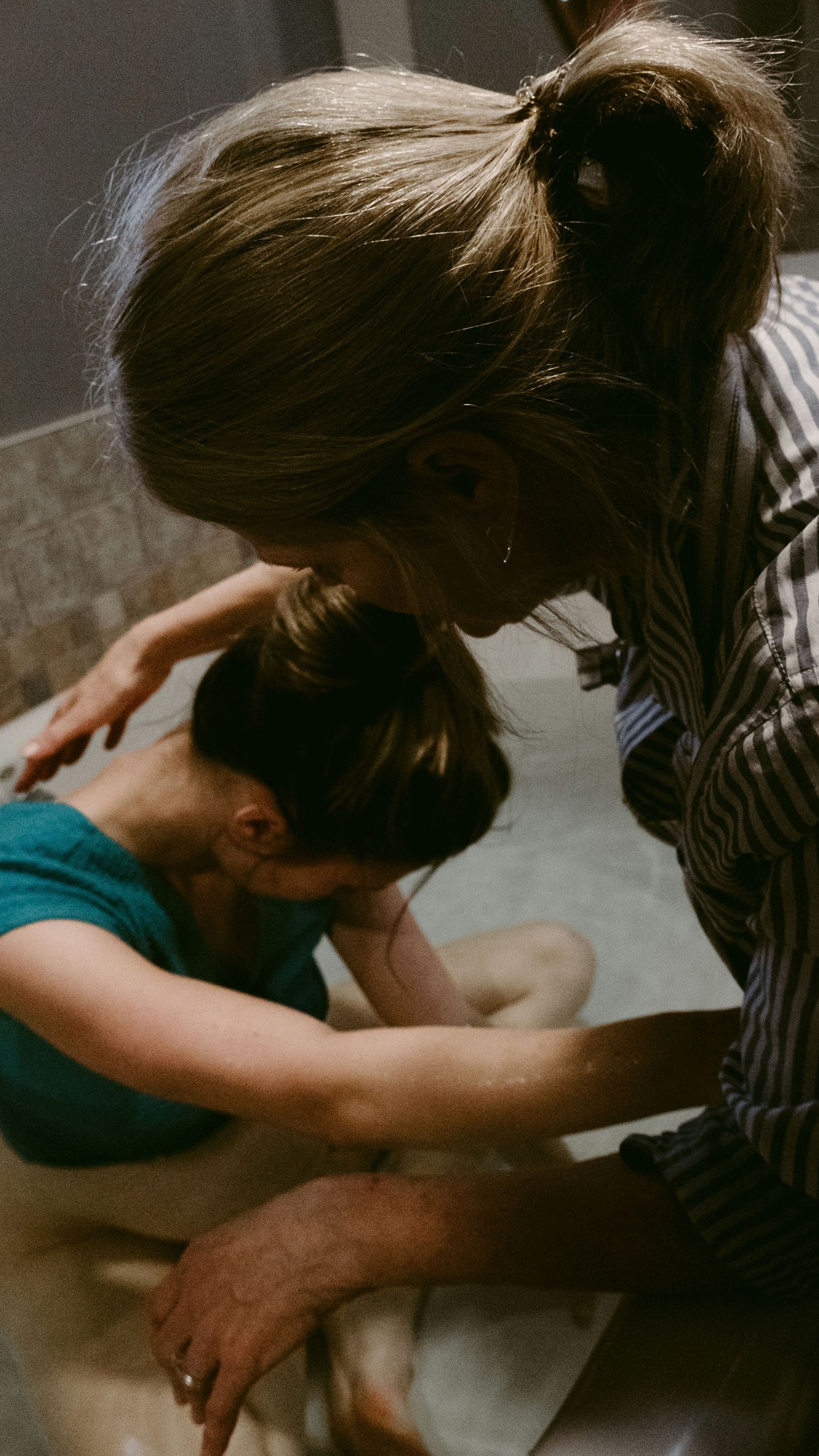 A woman in a striped shirt gently supports another person leaning over a bathtub in a bathroom setting.