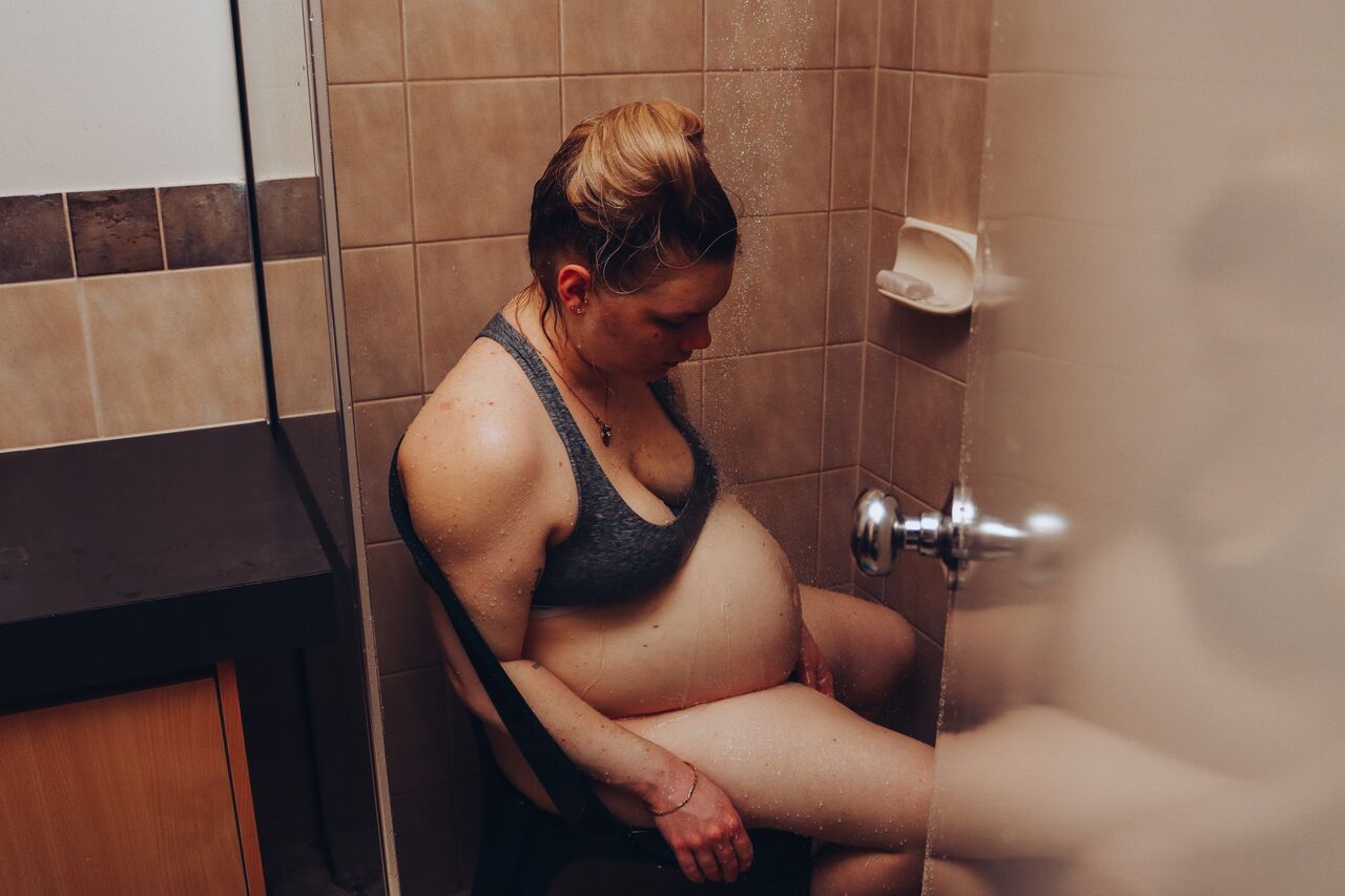 Pregnant woman in sports bra sitting on bathroom floor during labor, leaning against shower wall.
