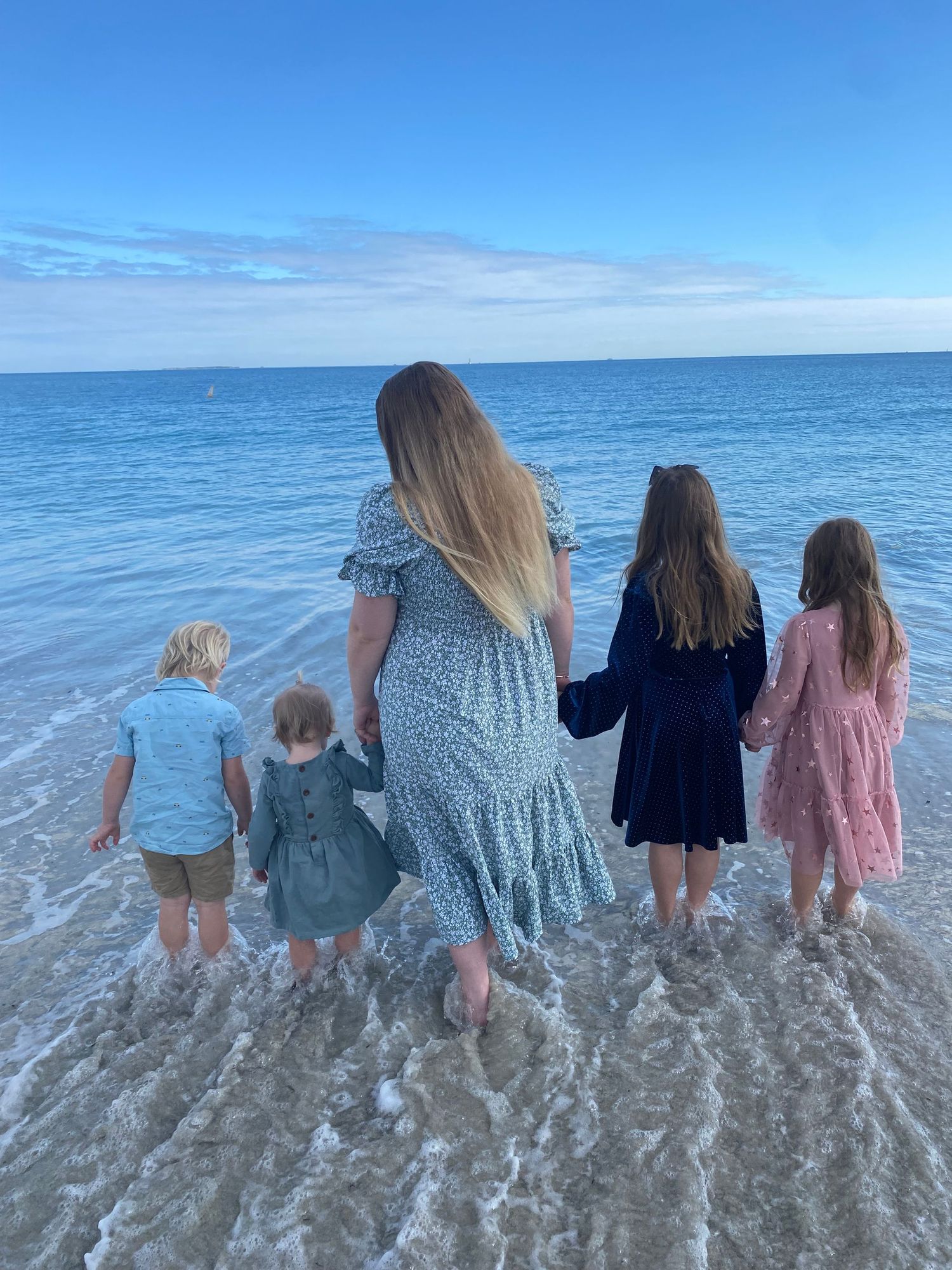 Mother with long blonde hair holding hands with four children walking toward ocean waves on beach.