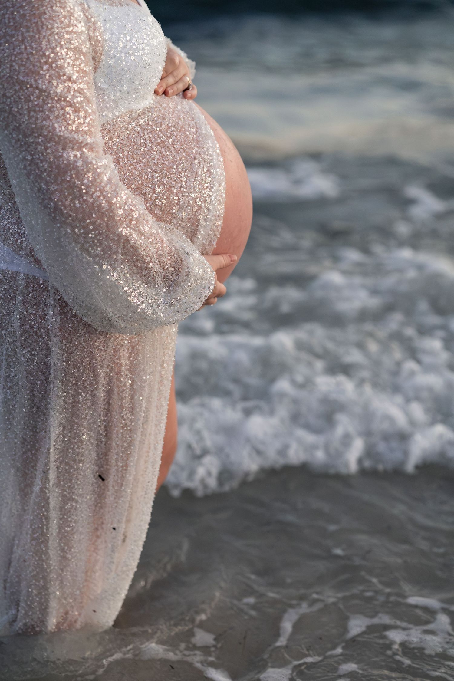 Pregnant woman in sheer white gown cradling baby bump while standing in ocean waves on beach.