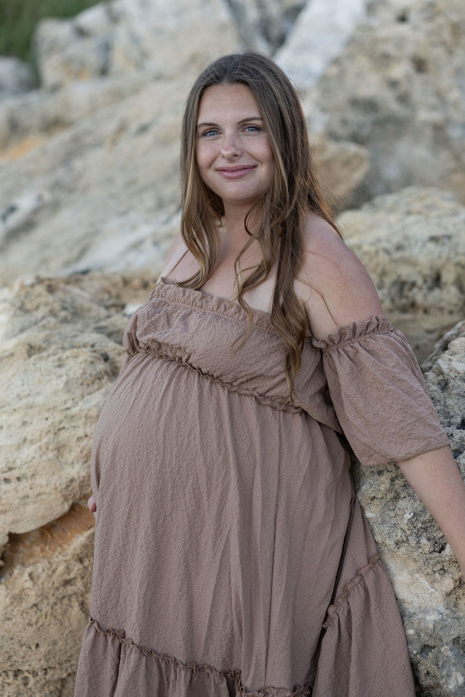 Pregnant woman in off-shoulder tan maxi dress smiling outdoors near rocky background during maternity photoshoot. Woodman Point Perth