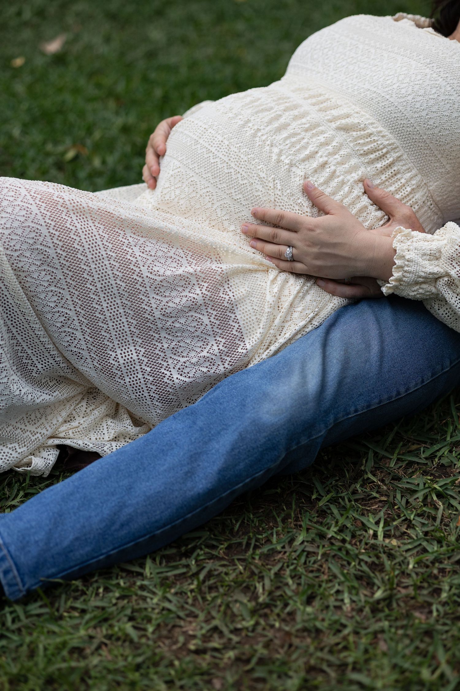 Pregnant woman in cream lace top and jeans sitting on grass, cradling her baby bump with both hands outdoors.  Araluen Perth