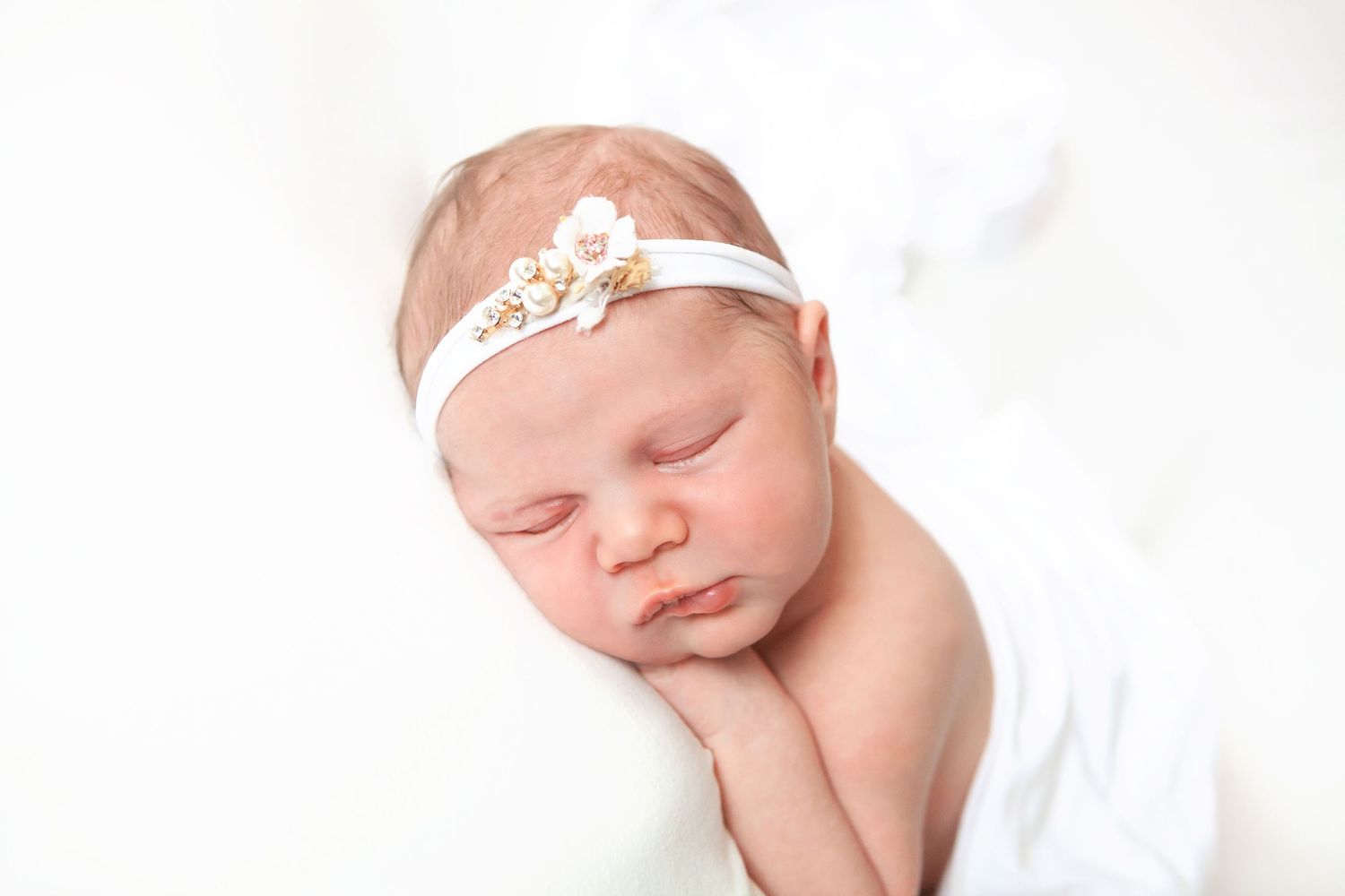 Sleeping newborn baby girl wearing a white floral headband, wrapped in a soft white blanket.