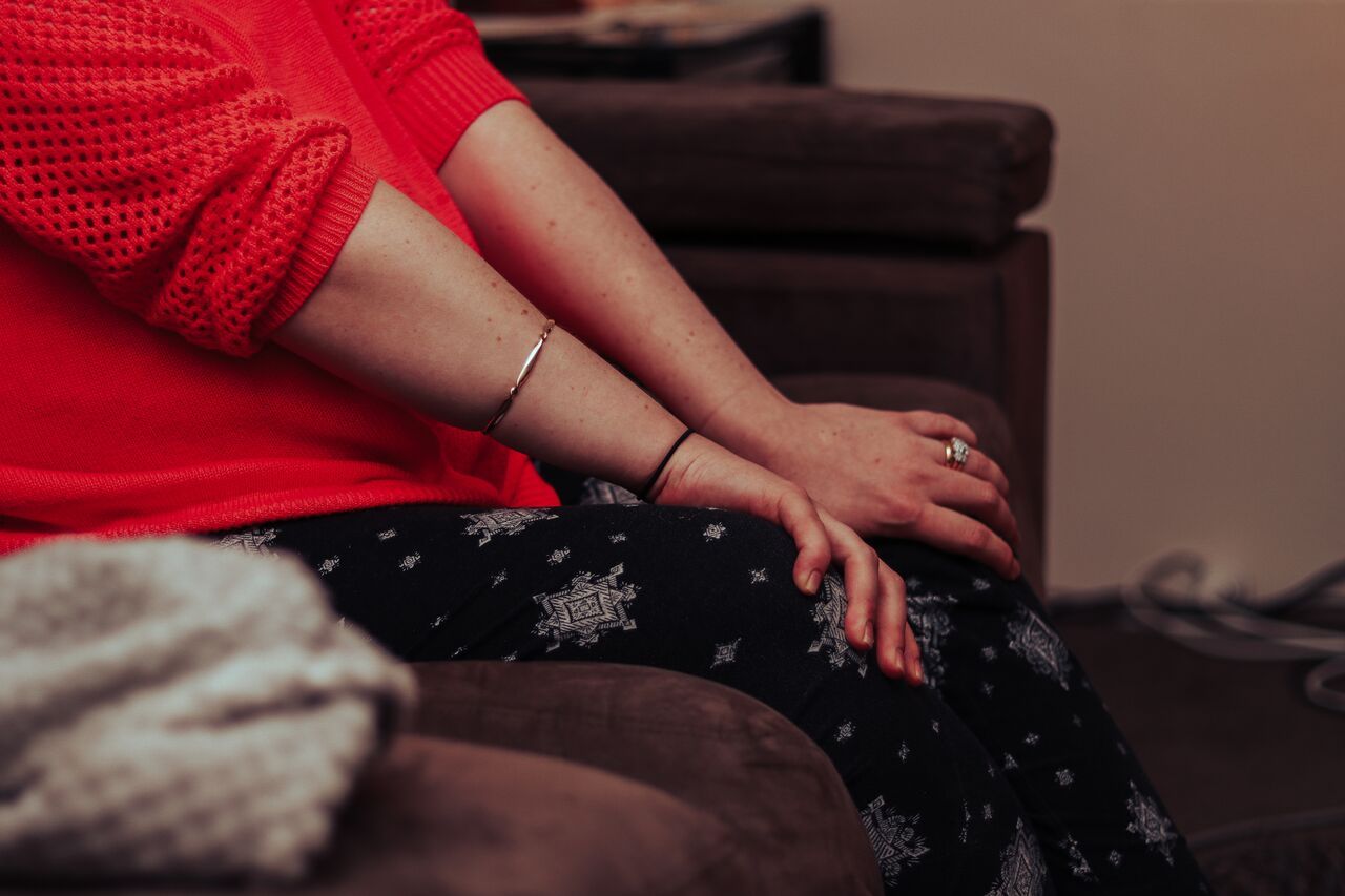 Woman in red sweater and snowflake leggings sitting on brown sofa with hands resting on lap.