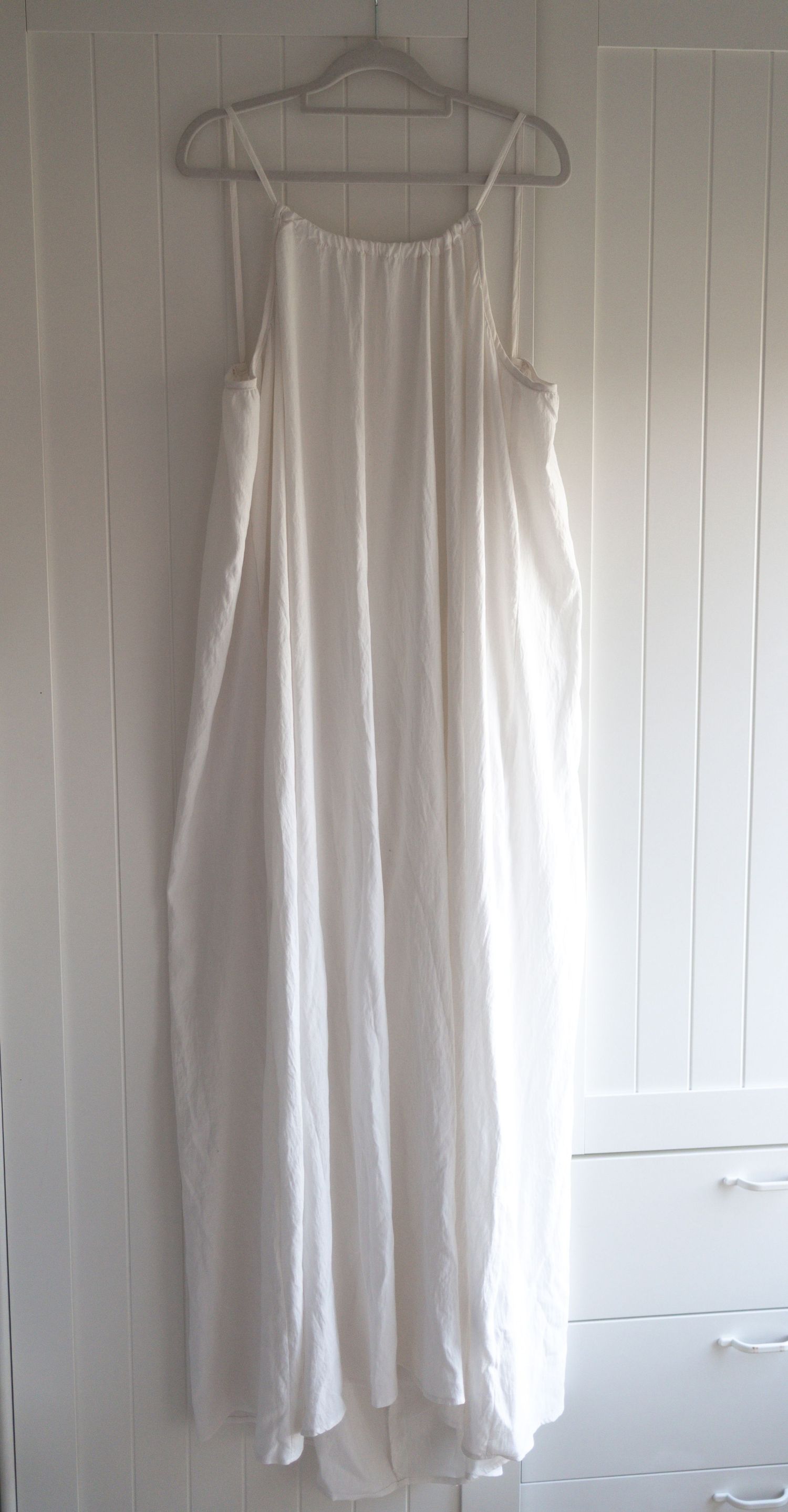 White maxi dress with spaghetti straps and relaxed silhouette hanging on a hanger against a white wall.