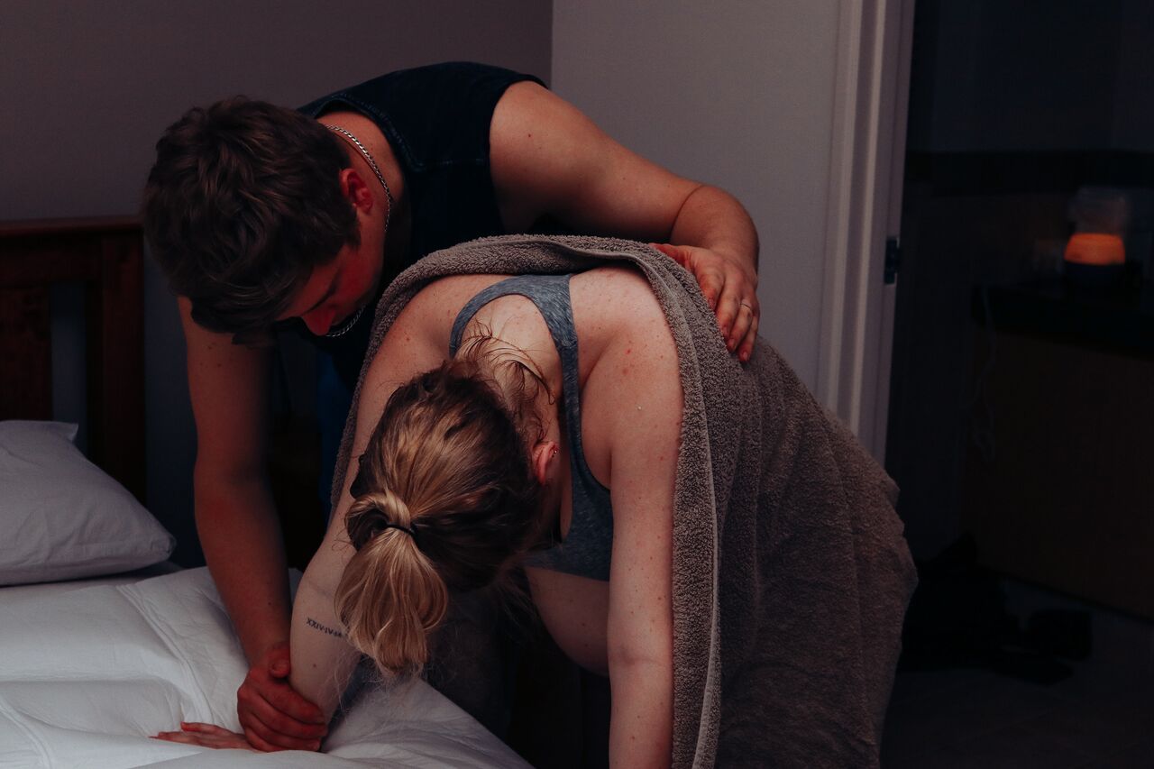 Partner providing back support to a laboring woman leaning over a bed in dim lighting.