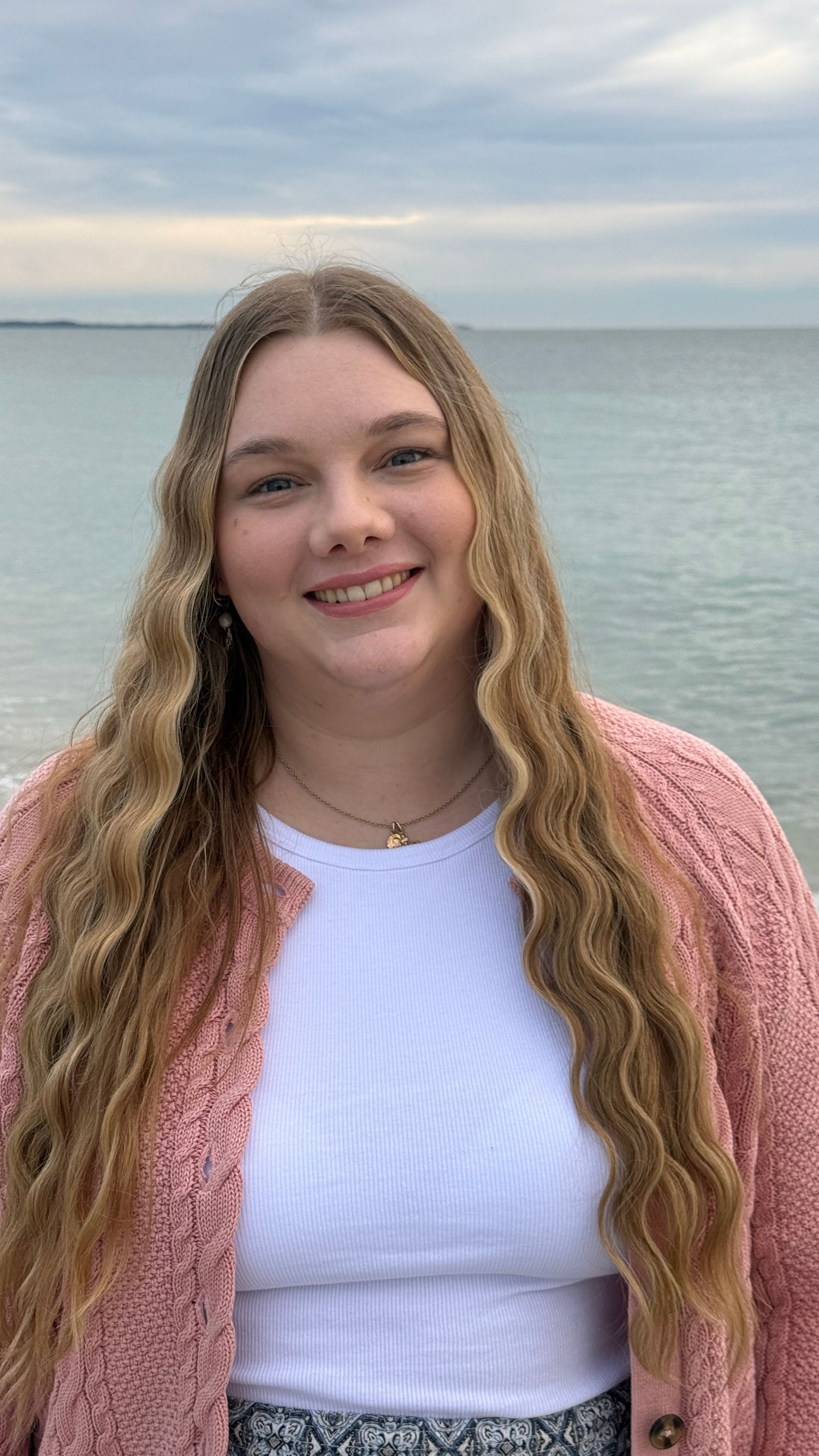 Smiling young woman with long wavy blonde hair wearing pink cardigan standing near ocean shoreline.