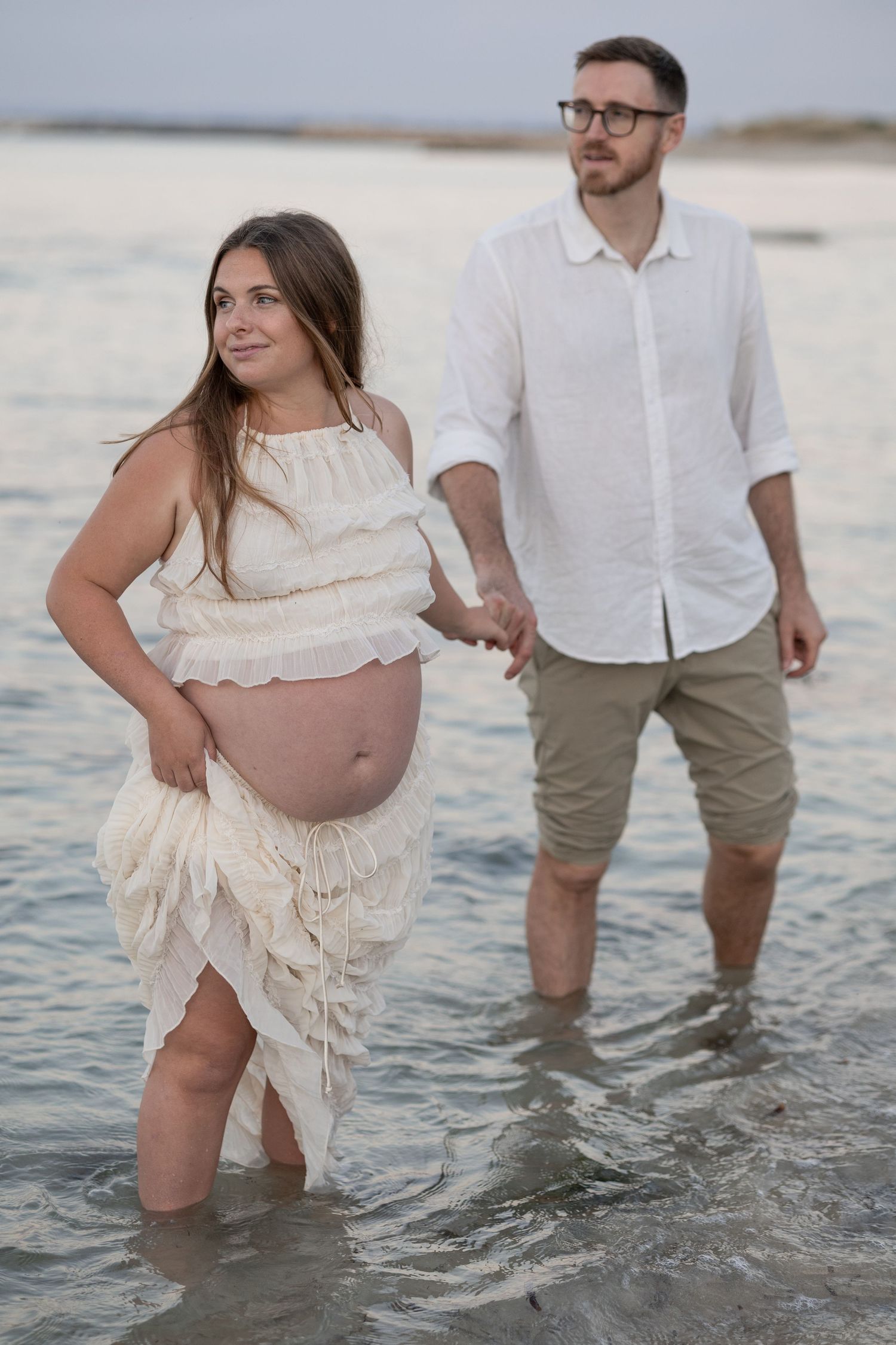 Pregnant couple holding hands standing in shallow ocean water, woman in white dress showing baby bump, man in white shirt.