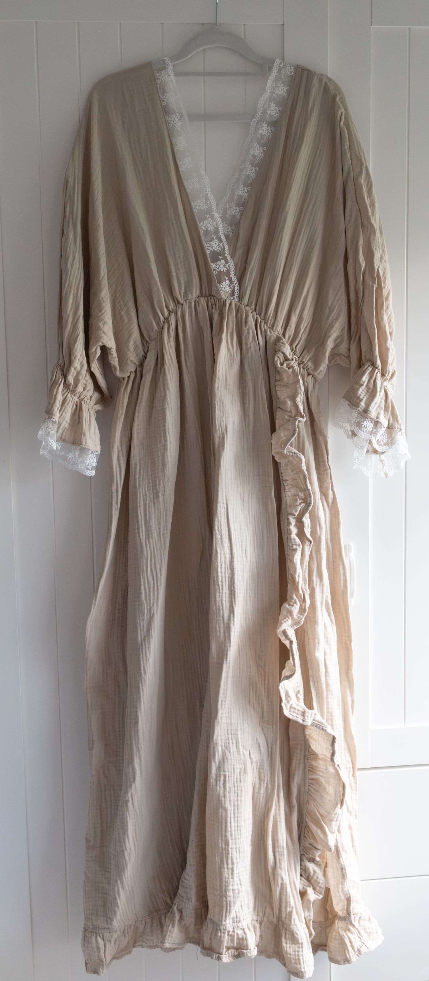 Boho beige maxi dress with lace trim, deep V-neck, ruffled hem, and bell sleeves, hanging on white wall.