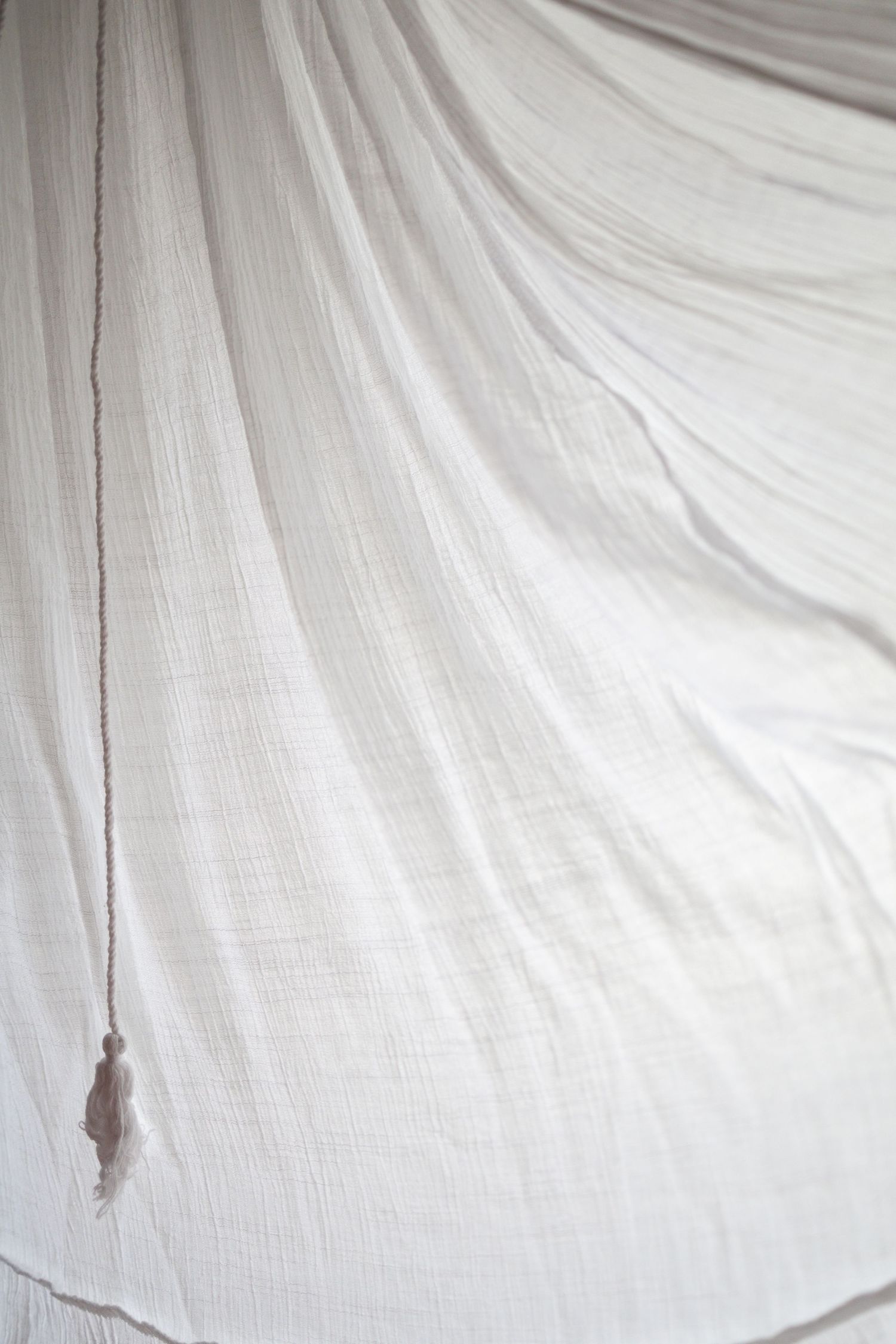 Soft white draped muslin fabric with elegant folds and a delicate tassel detail, creating a serene, airy backdrop.
