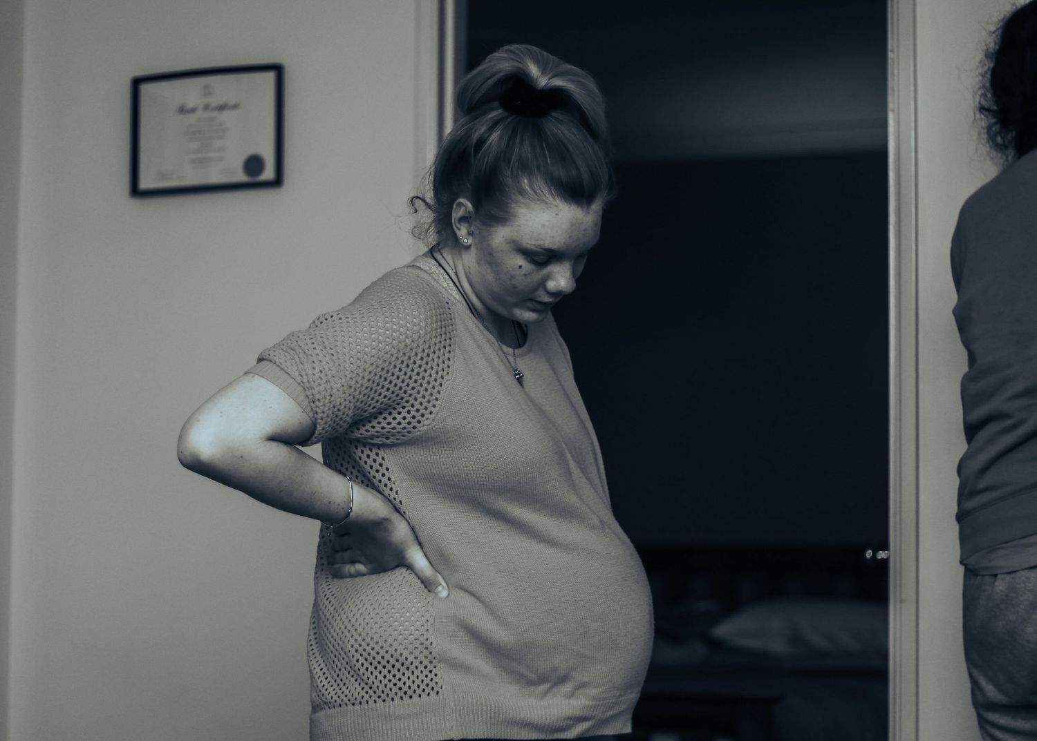 Pregnant woman holding her lower back in pain, standing in a room in a black and white photo.