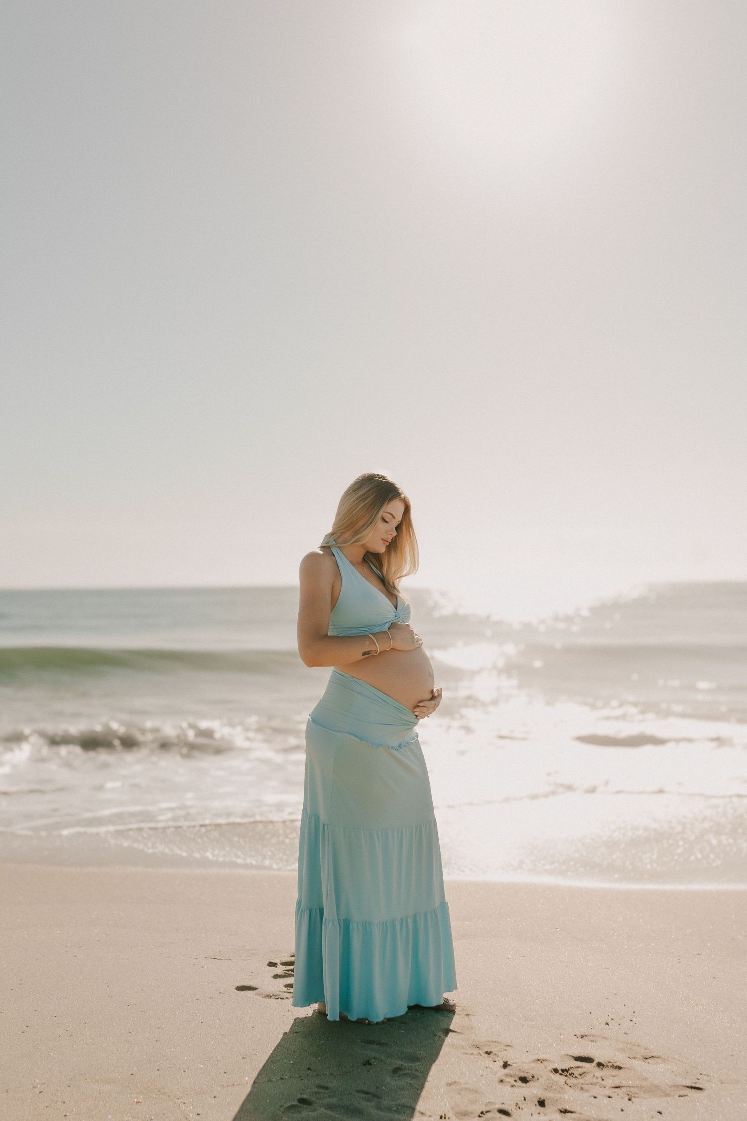Pregnant woman in light blue maxi dress cradling her baby bump on a sunny beach with ocean waves behind her.