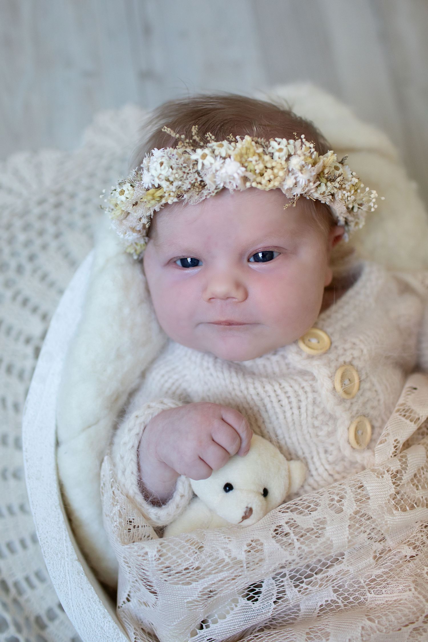 Newborn baby wearing floral crown and cream knit outfit, holding small white teddy bear in lace-lined basket.