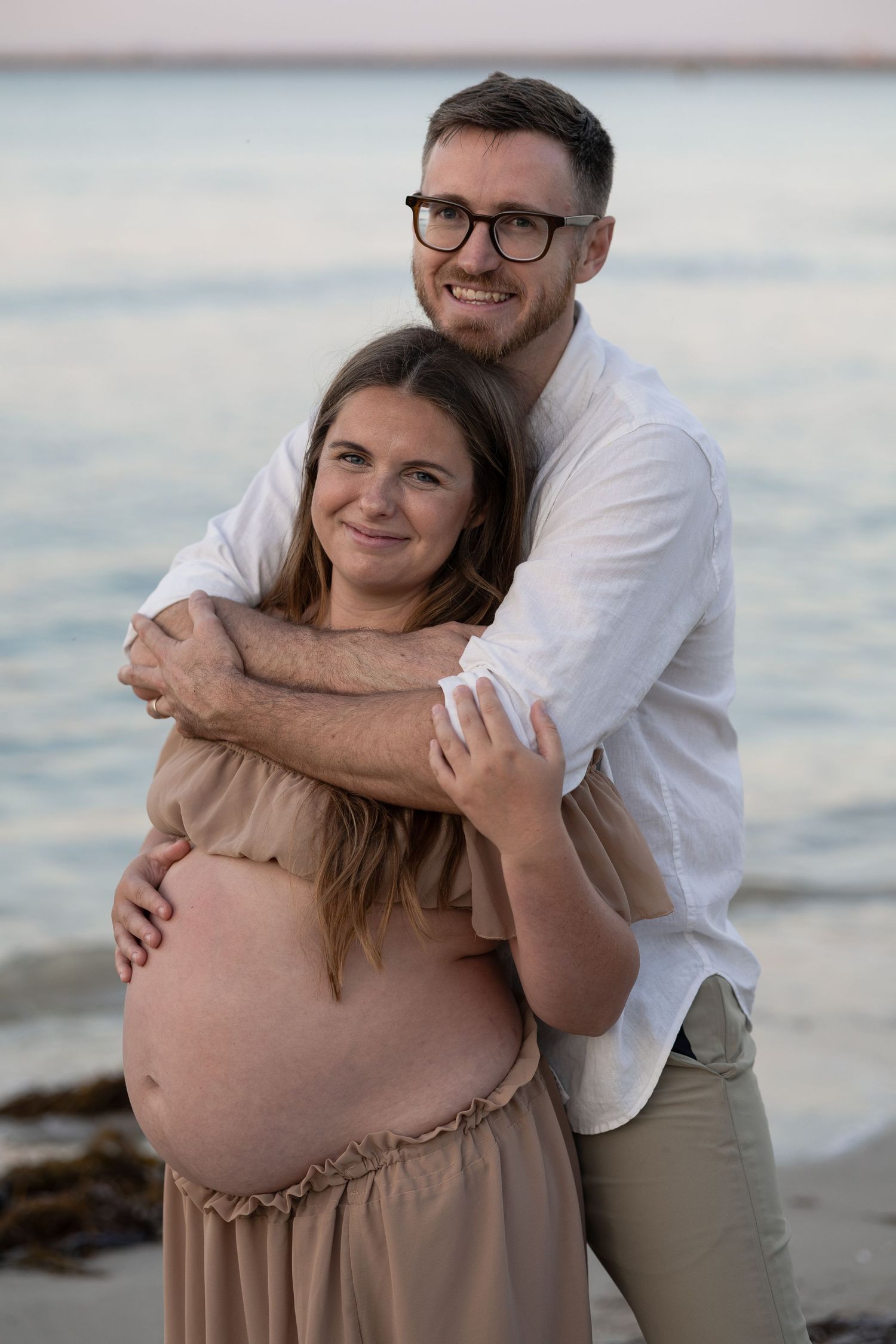 Smiling couple embracing on beach, pregnant woman in tan dress with baby bump visible, man in white shirt and glasses.