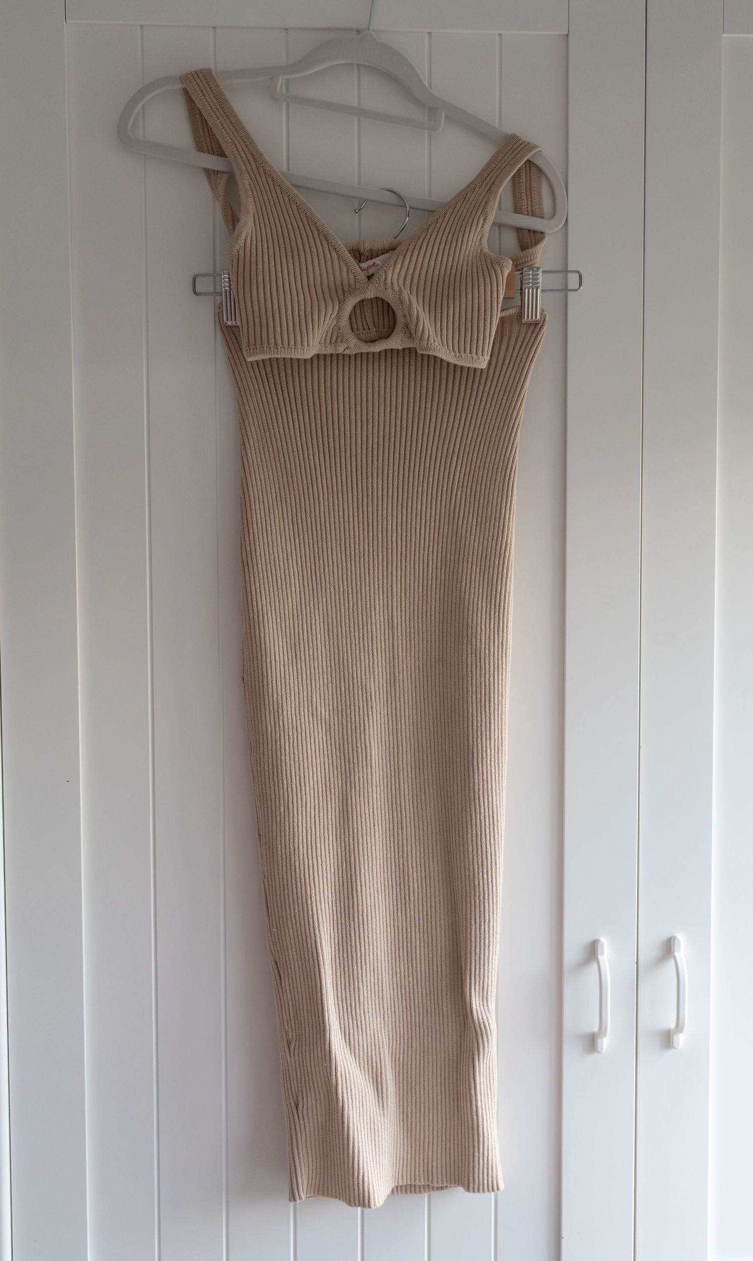 Beige ribbed knit midi dress with cut-out bust detail and O-ring hardware, displayed on hanger against white background.